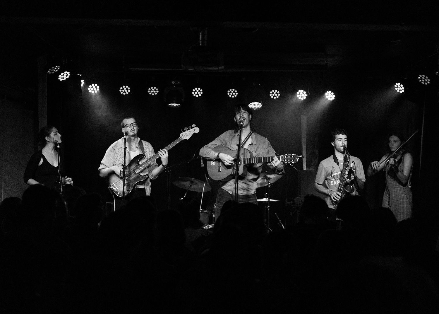 Black and white photo of a five-piece band performing live on a lit stage with guitars and microphones.