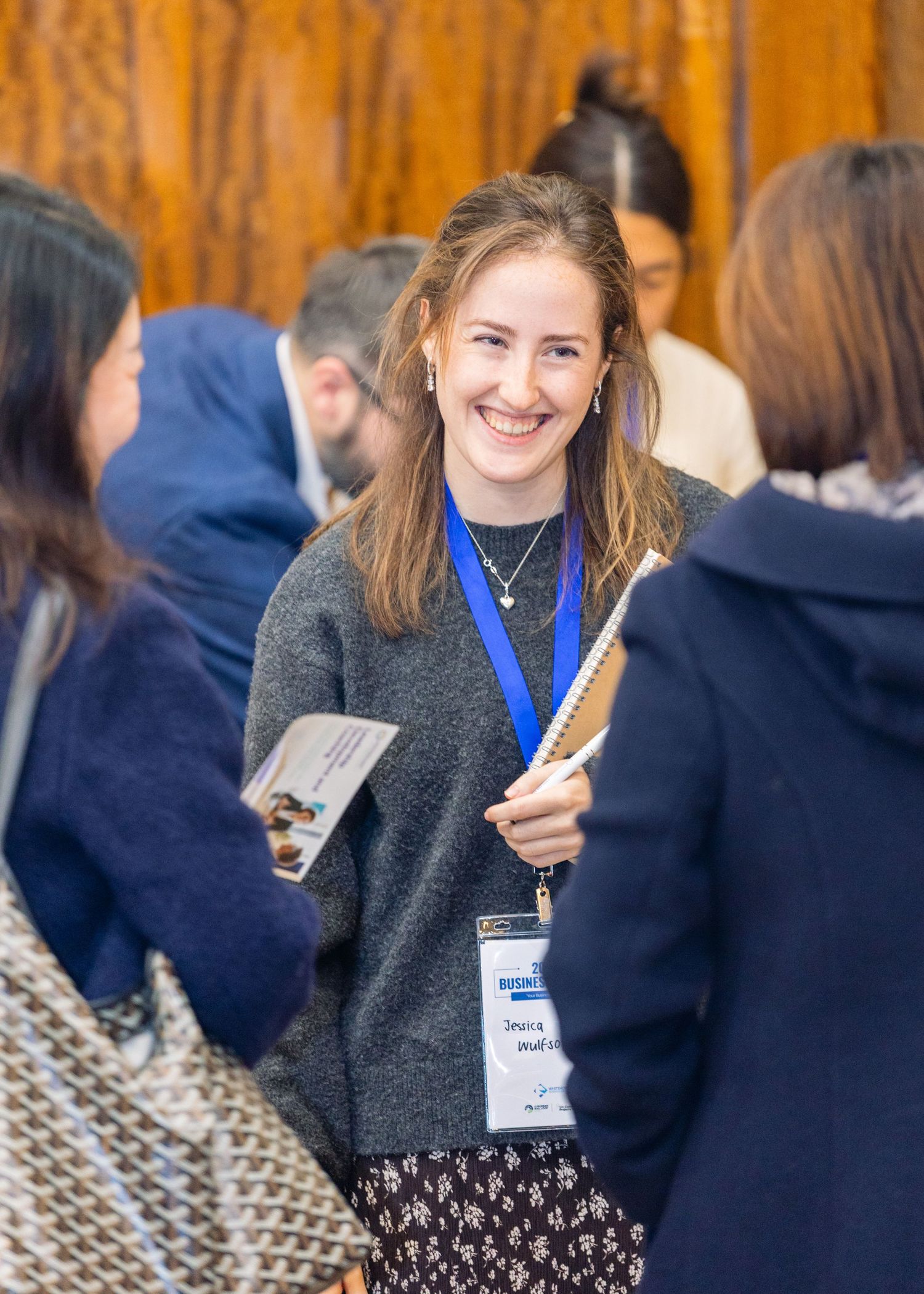 Smiling young woman wearing a blue lanyard and holding a notepad at a professional networking conference event.