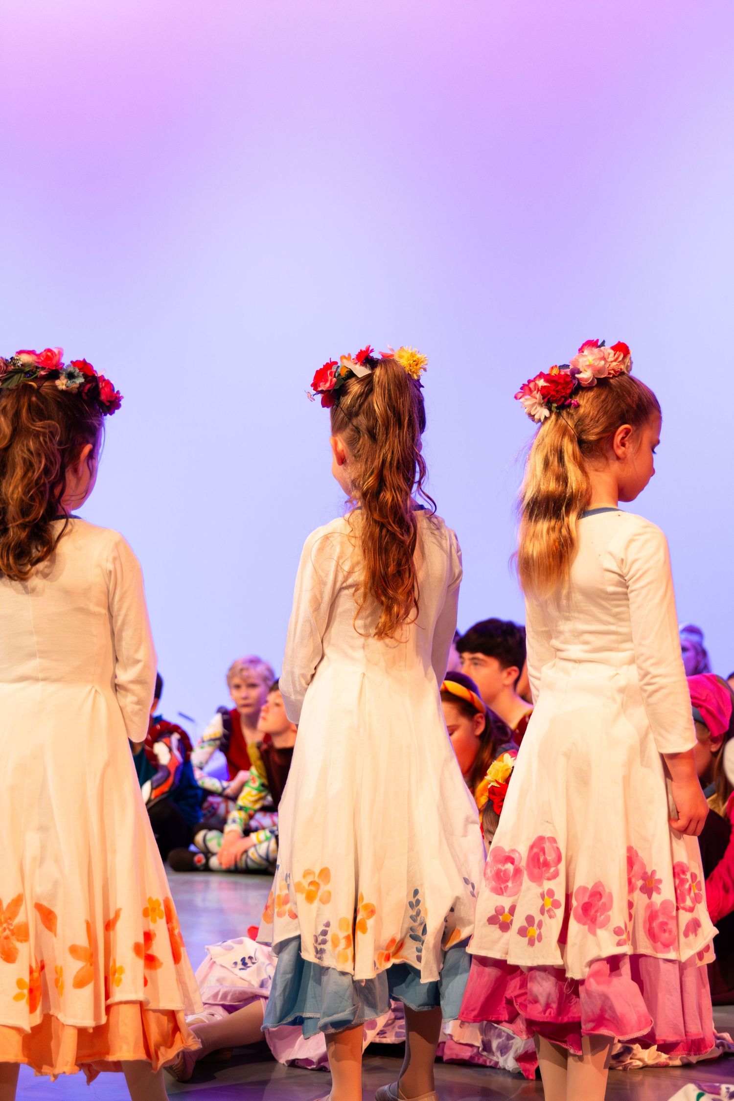 Three girls in white floral dresses with flower crowns stand on stage during a performance.