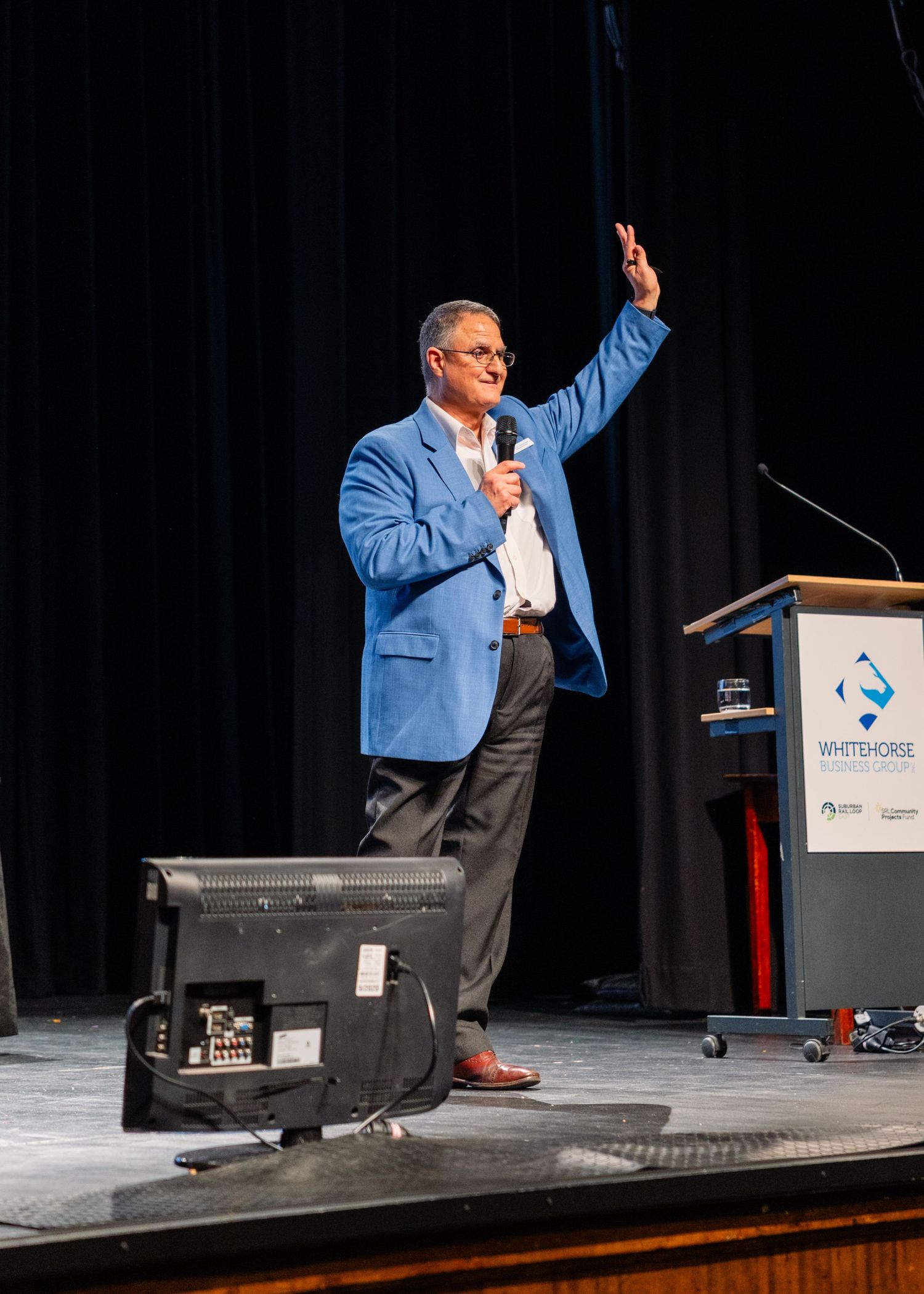 Male speaker in blue blazer presenting on stage with microphone at Whitehorse conference event.