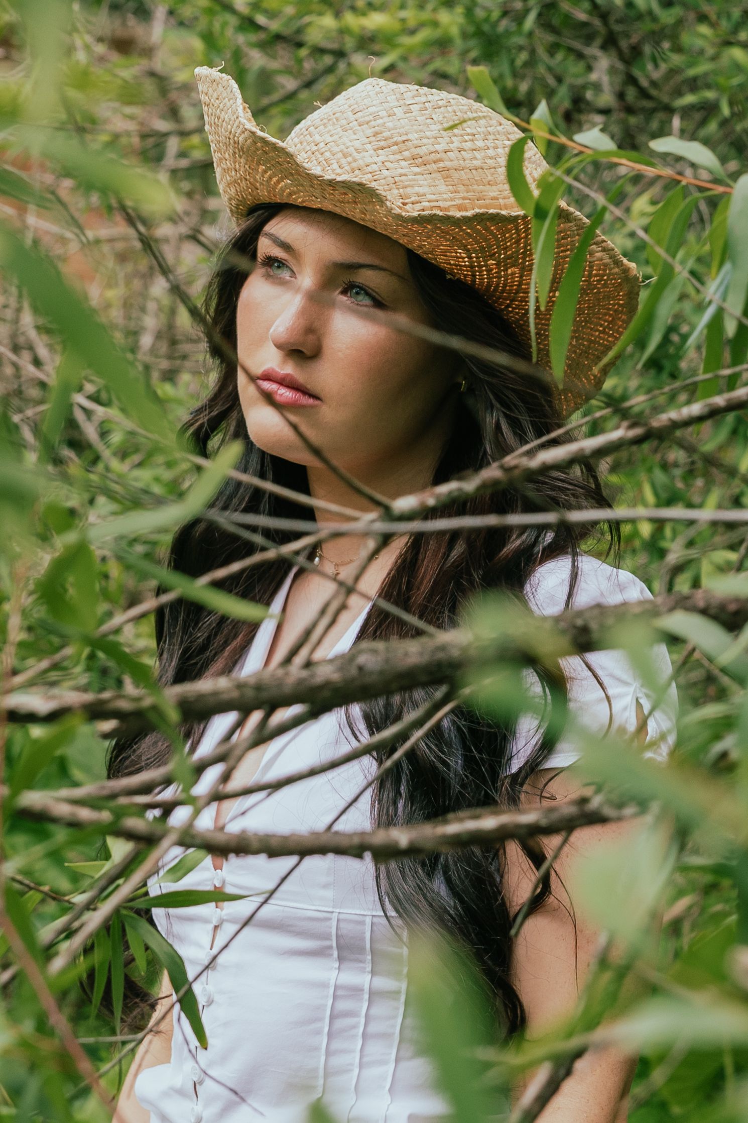 Young woman in straw cowboy hat and white dress posed among green leafy branches outdoors.