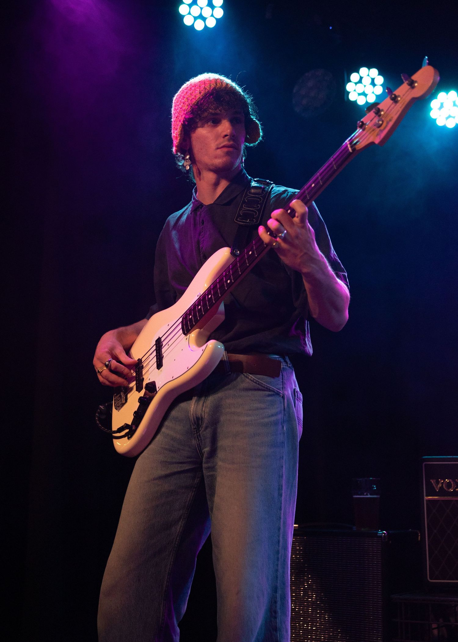 Young male guitarist in a red beanie playing a white electric bass guitar on a colorfully lit stage.