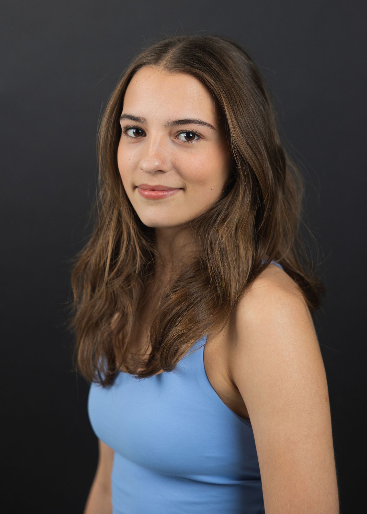 Young woman with brown hair wearing a light blue top, smiling against a dark gray background.