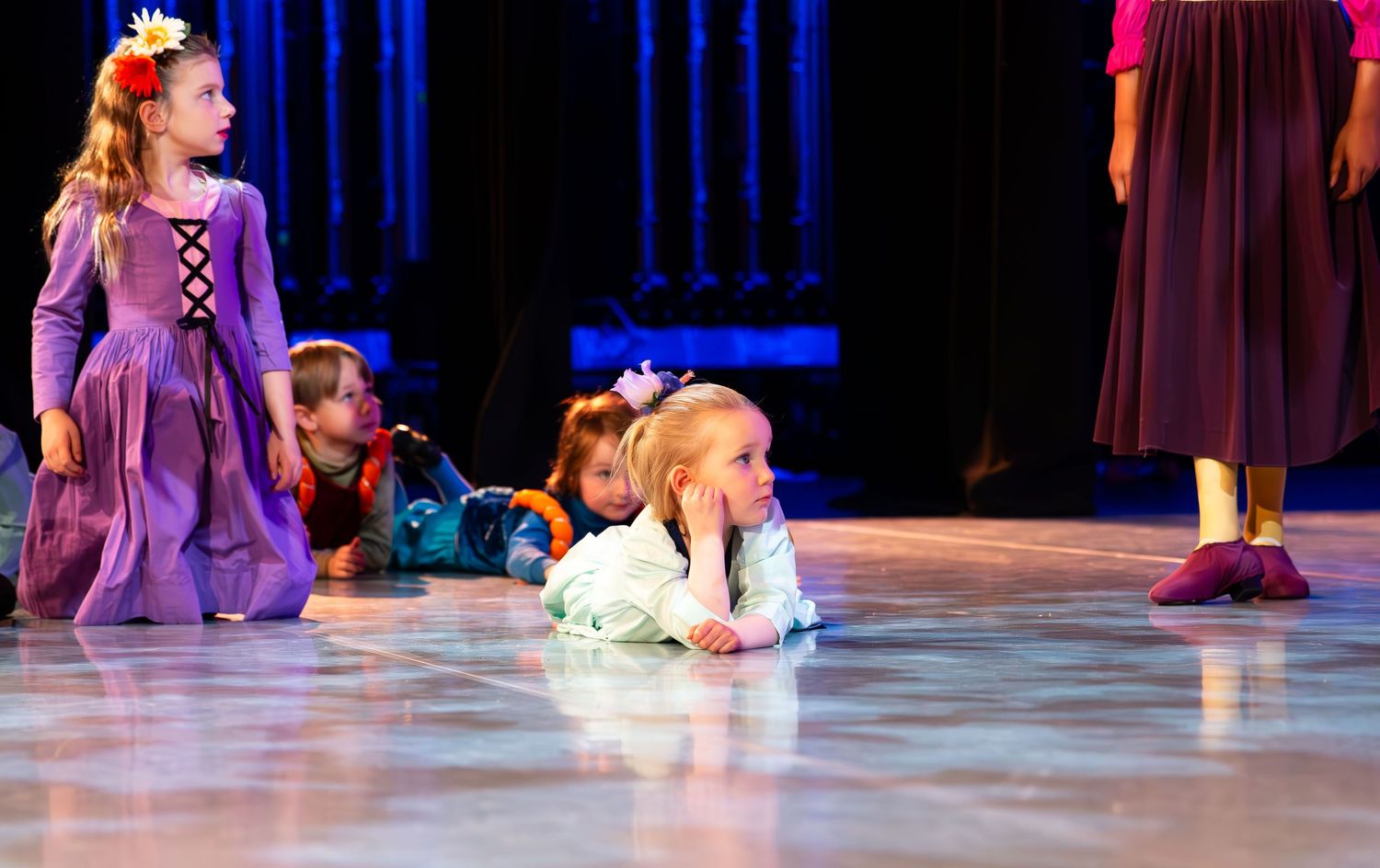 Young girls in costumes perform on stage, with one crawling on the floor during a children's theater production.