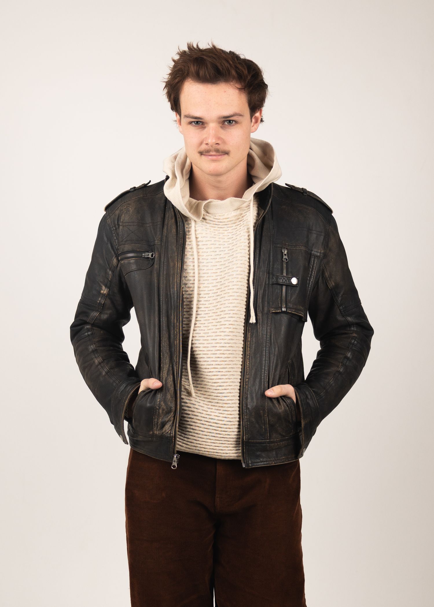 Young man wearing a distressed black leather jacket over a cream knit hoodie sweater with brown corduroy pants.