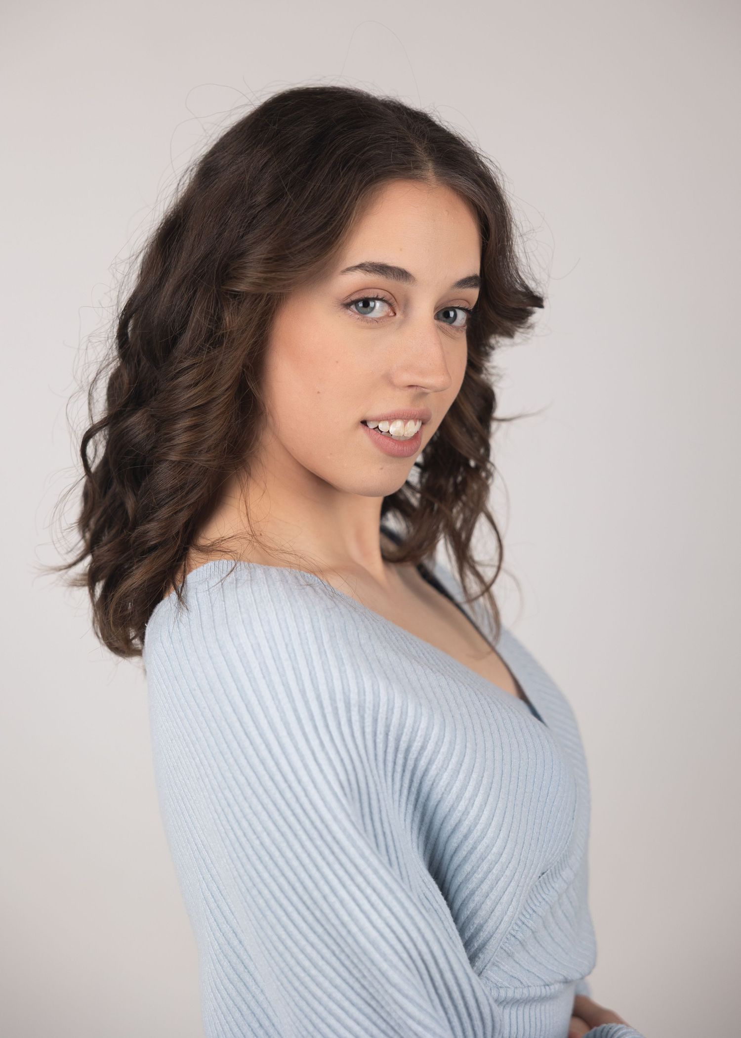 Young woman with dark curly hair in a light blue ribbed sweater smiling over her shoulder against a white background.