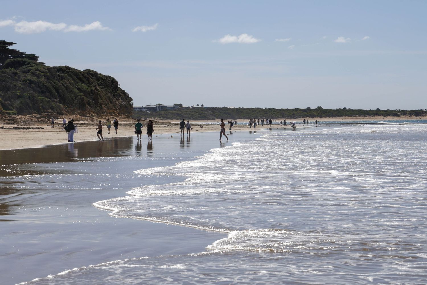 People walking along a scenic beach with gentle waves, sandy shores, and a rocky cliff under a partly cloudy sky.