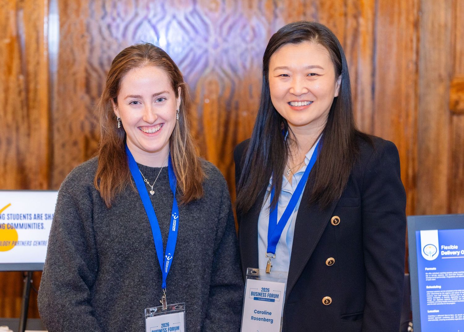 Two women wearing blue lanyards smile together at a professional conference event indoors.