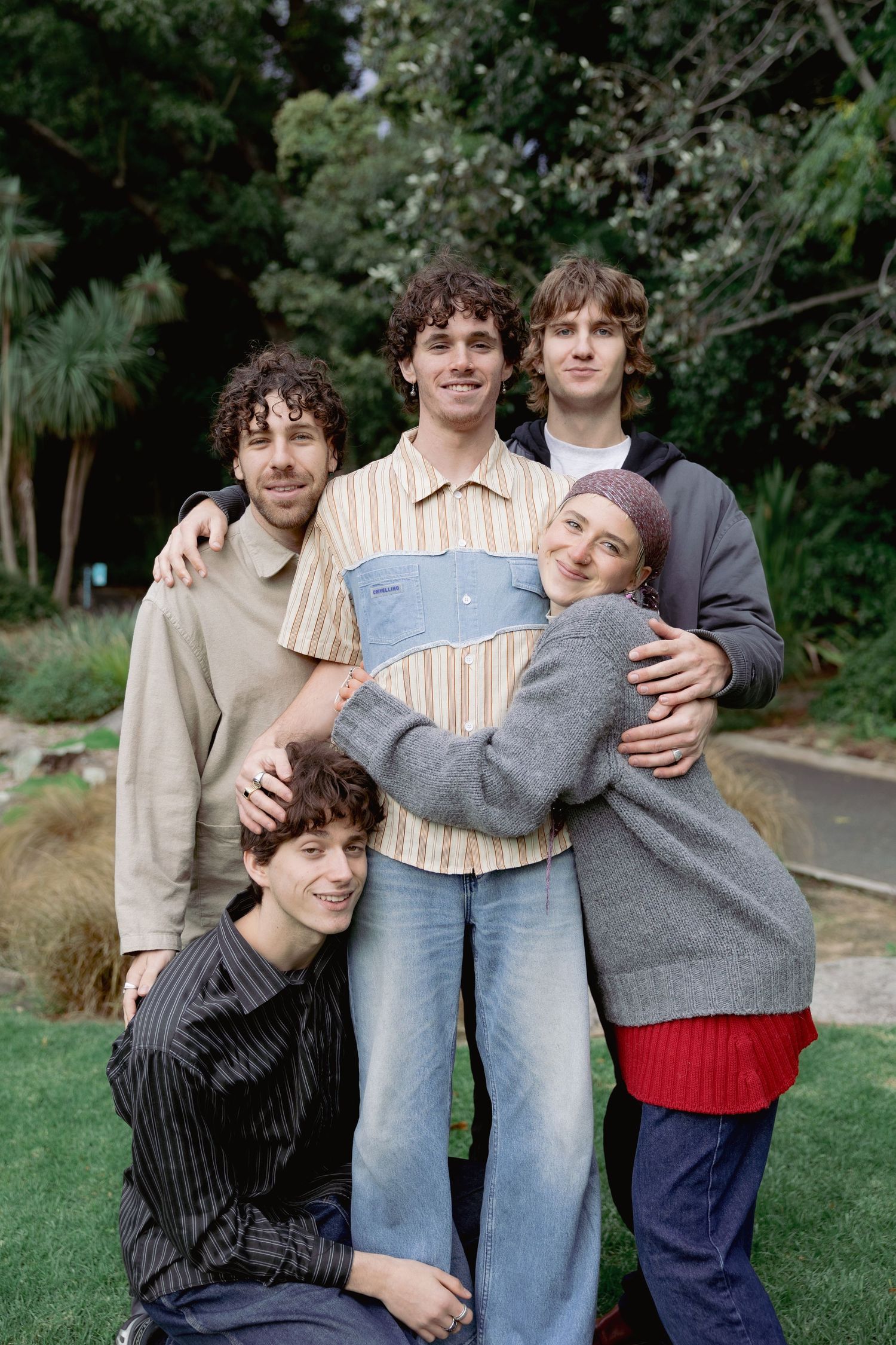 Five young friends pose together smiling in a lush green park, hugging closely for a group portrait.