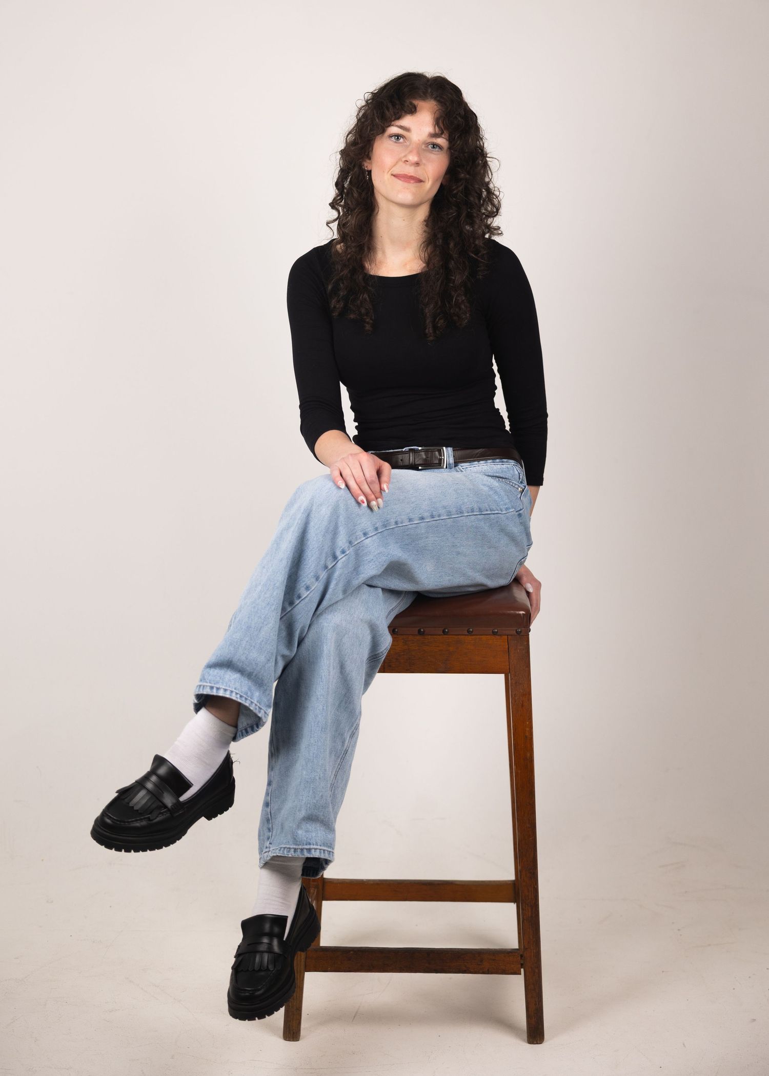 Woman with curly hair sitting on wooden stool wearing black top, light jeans, and black loafers.