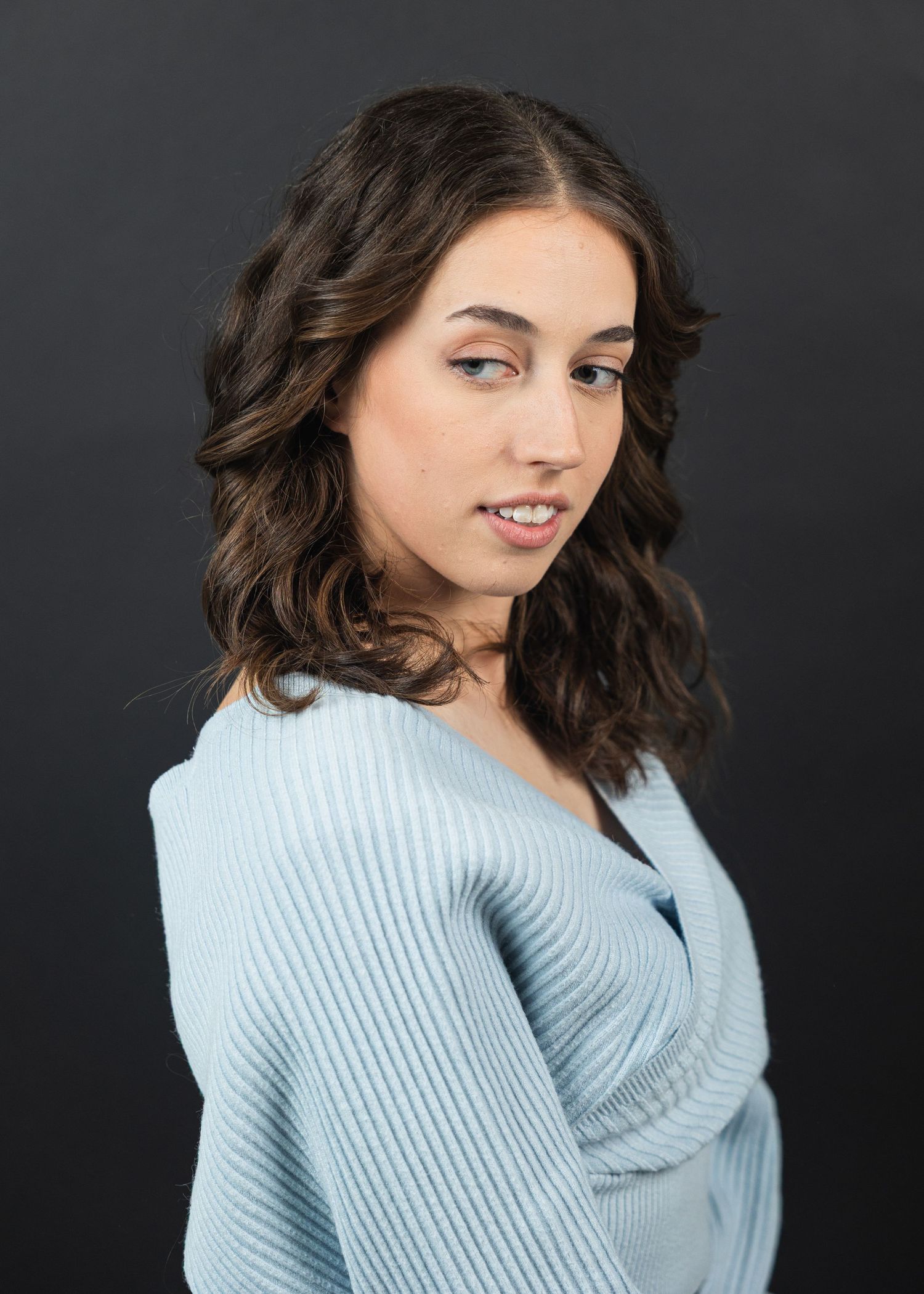 Young woman with wavy dark hair poses in a light blue ribbed sweater against a dark gray background.