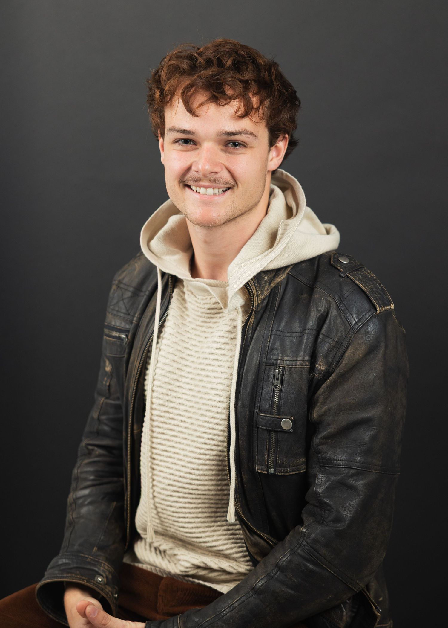 Young man smiling in a black leather jacket and cream hoodie against a dark background.