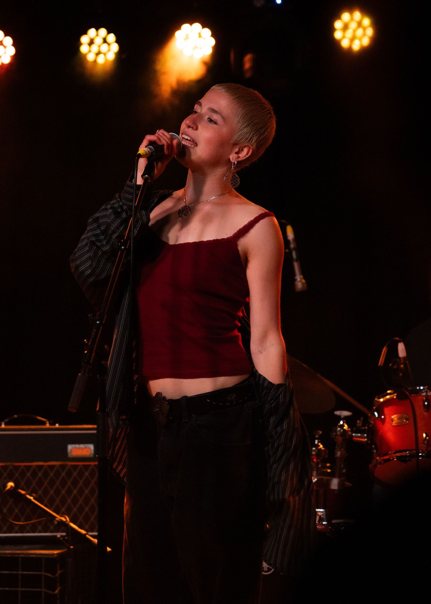 Young female singer performing on stage in red crop top under warm stage lights with band equipment visible.