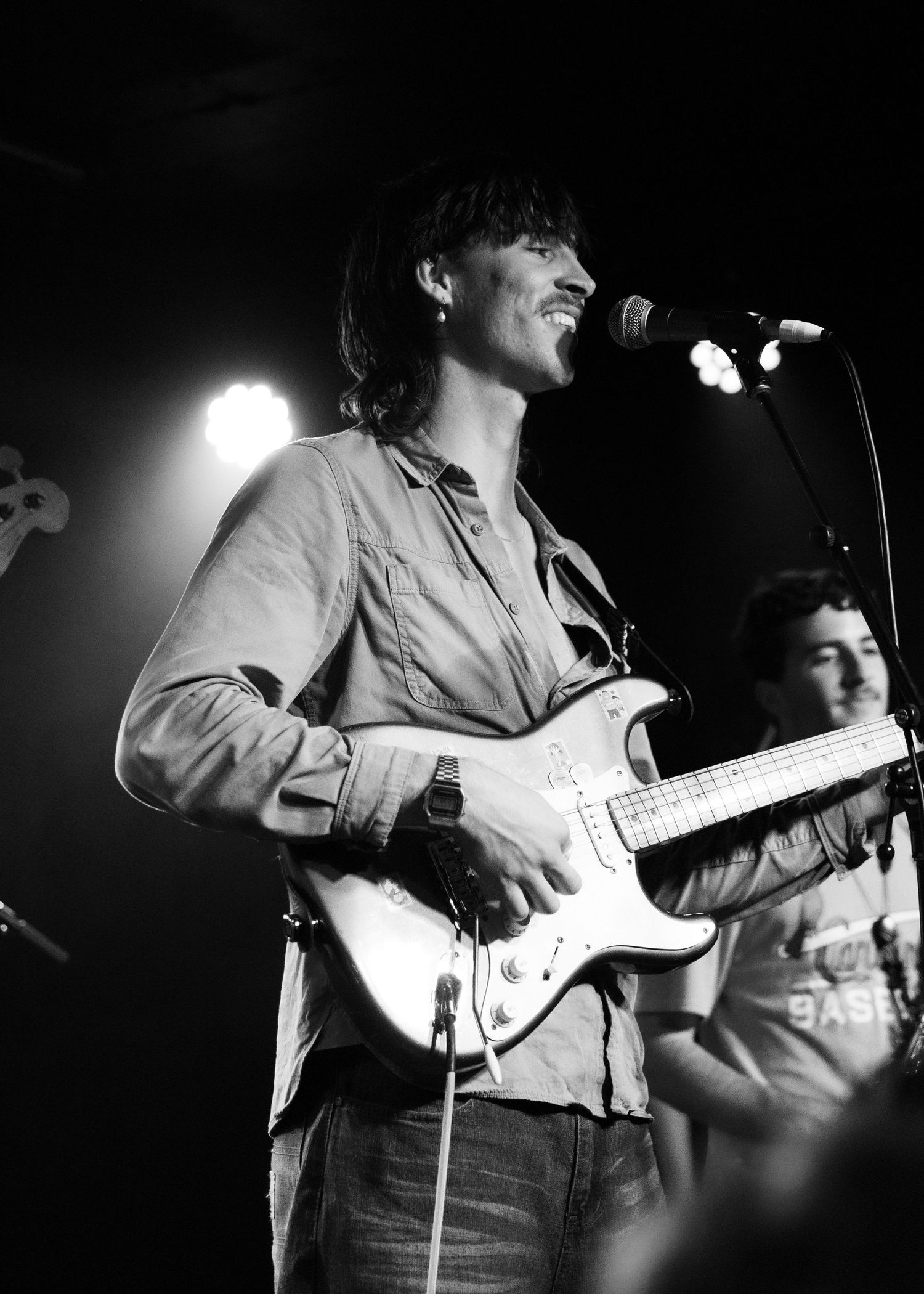 Smiling guitarist playing electric guitar at microphone on stage during a black and white live concert.