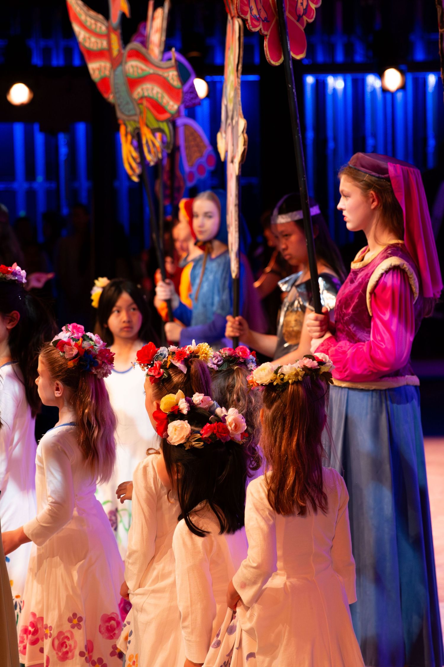Girls in floral crowns and colorful costumes perform on stage during a theatrical production.