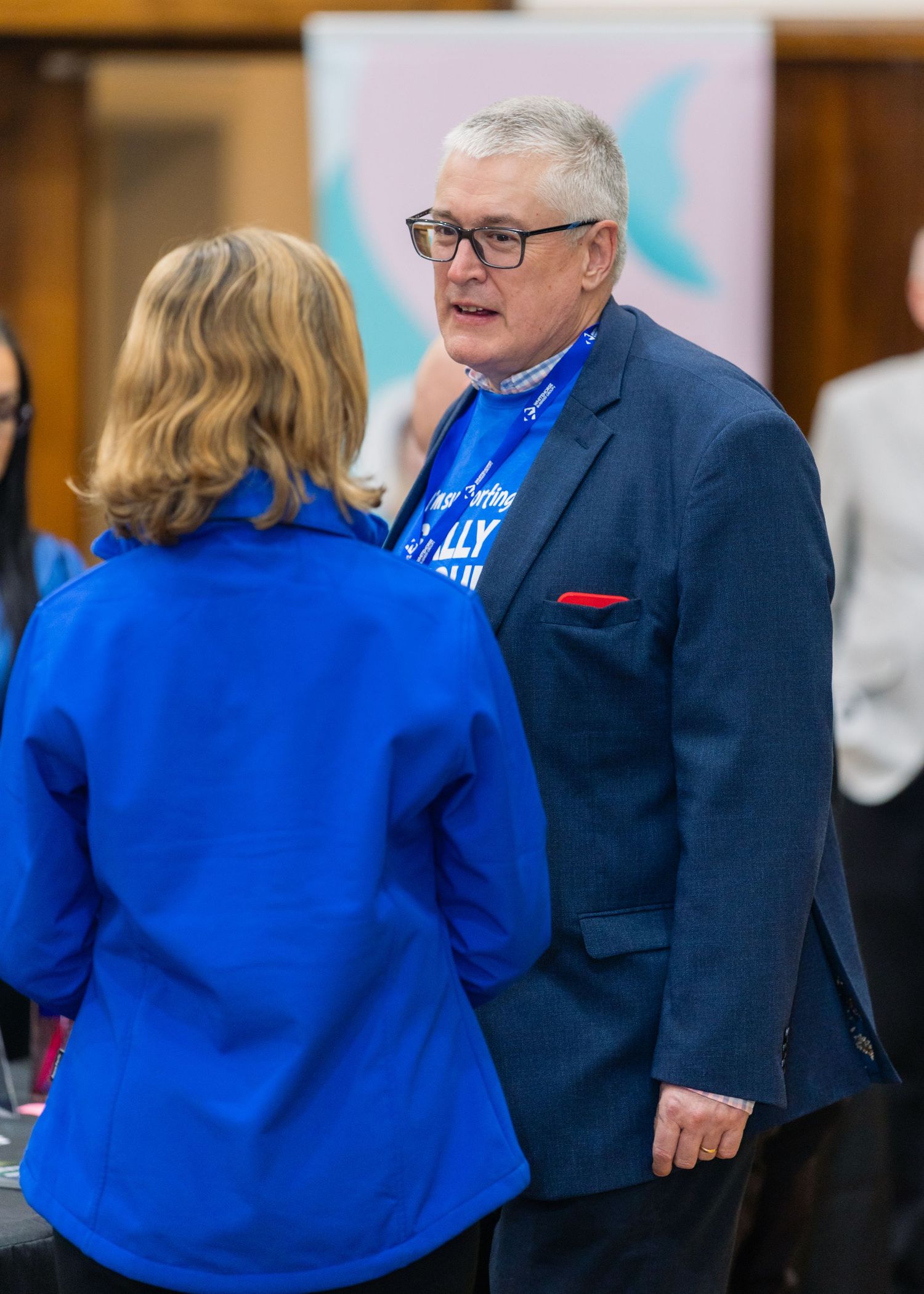 Man in blue blazer and glasses converses with woman in blue jacket at a professional networking event.