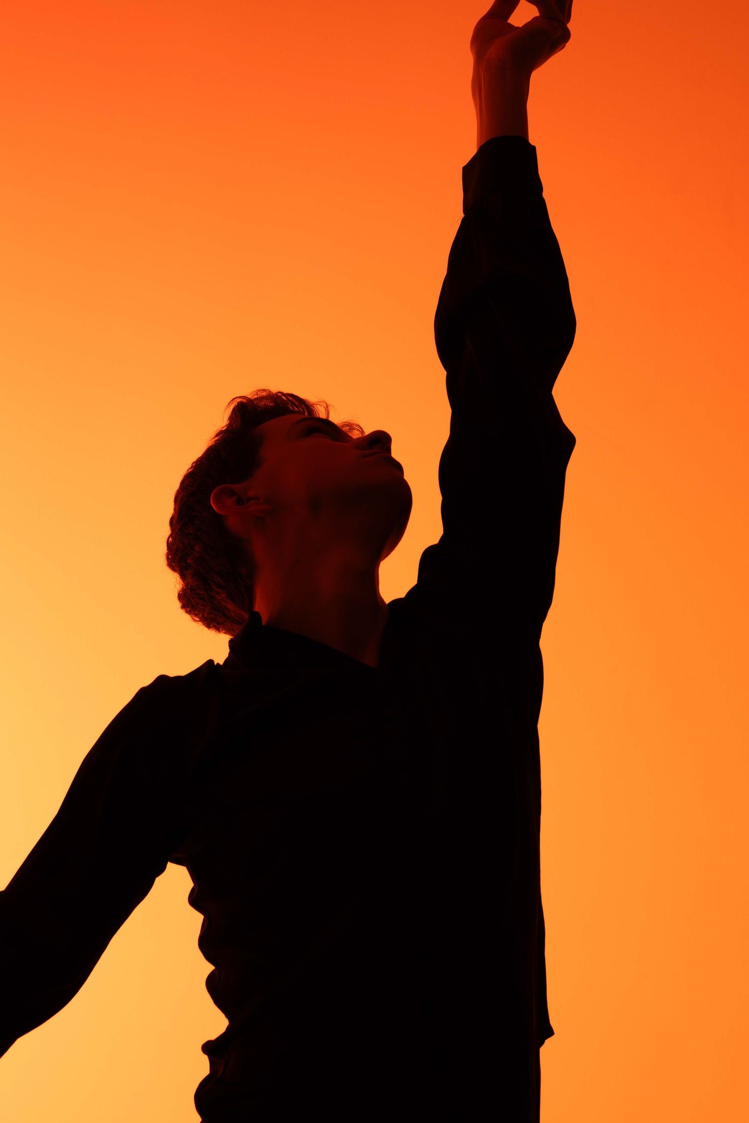Silhouette of person reaching upward dramatically against vibrant orange sunset sky background.