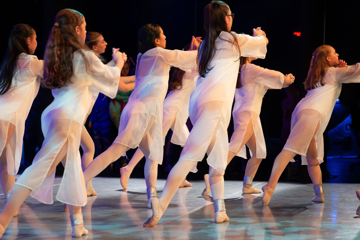 Group of young female dancers in white outfits performing synchronized routine on stage under colorful lights.
