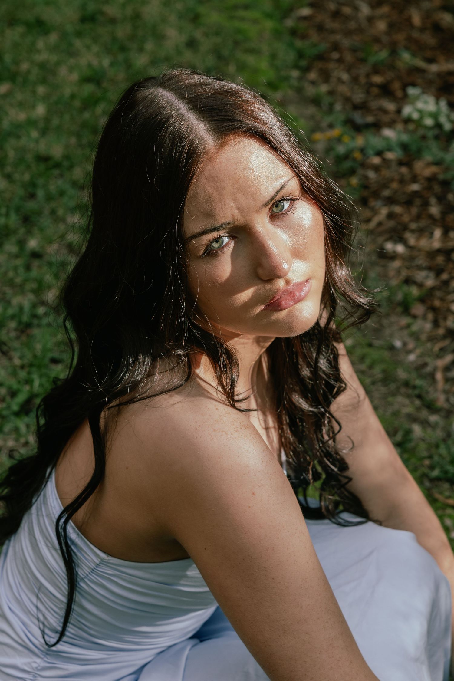 Young woman with dark wavy hair in white dress sitting outdoors on grass, looking at camera.