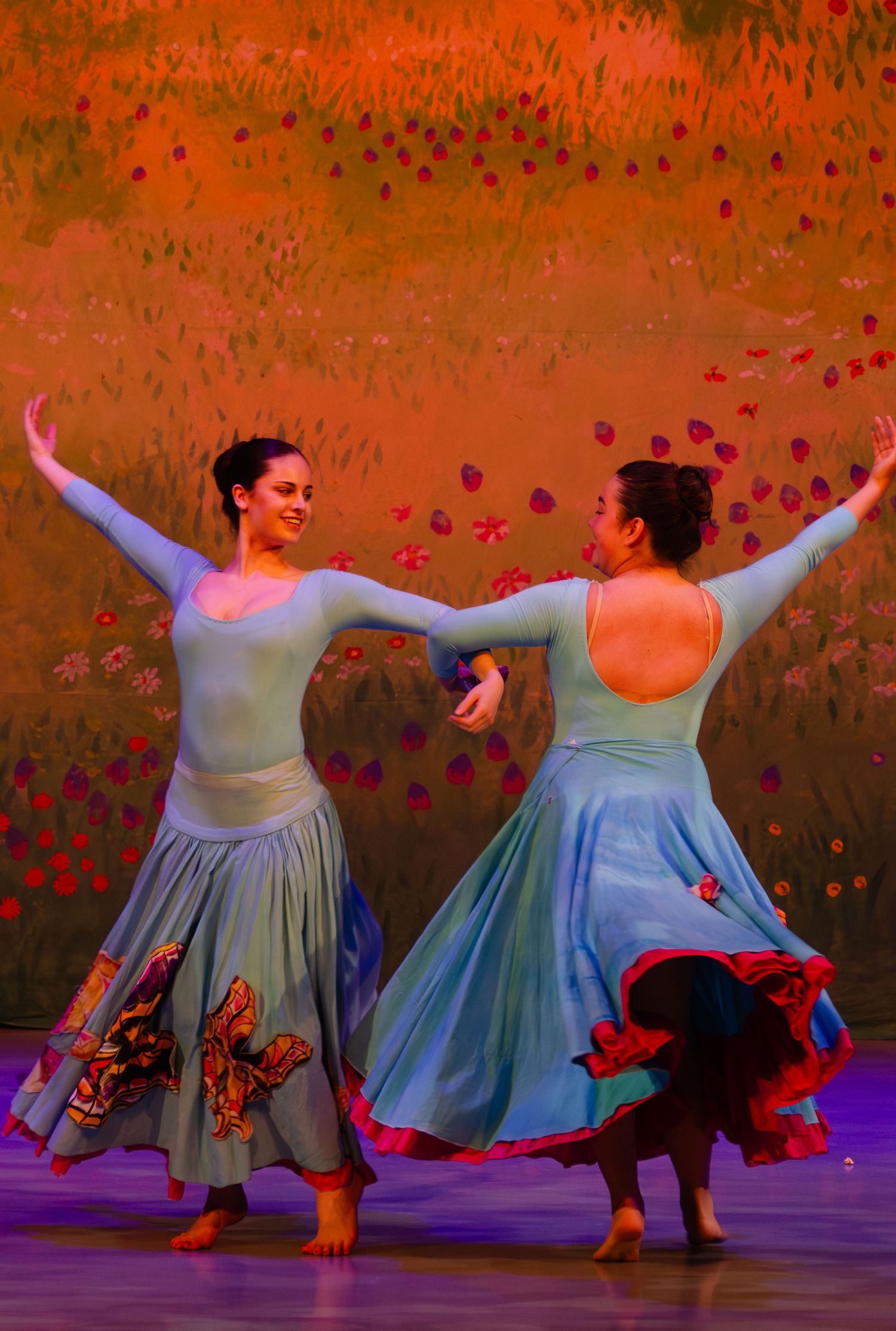 Two dancers in flowing blue dresses perform a graceful duet against a warm orange background.