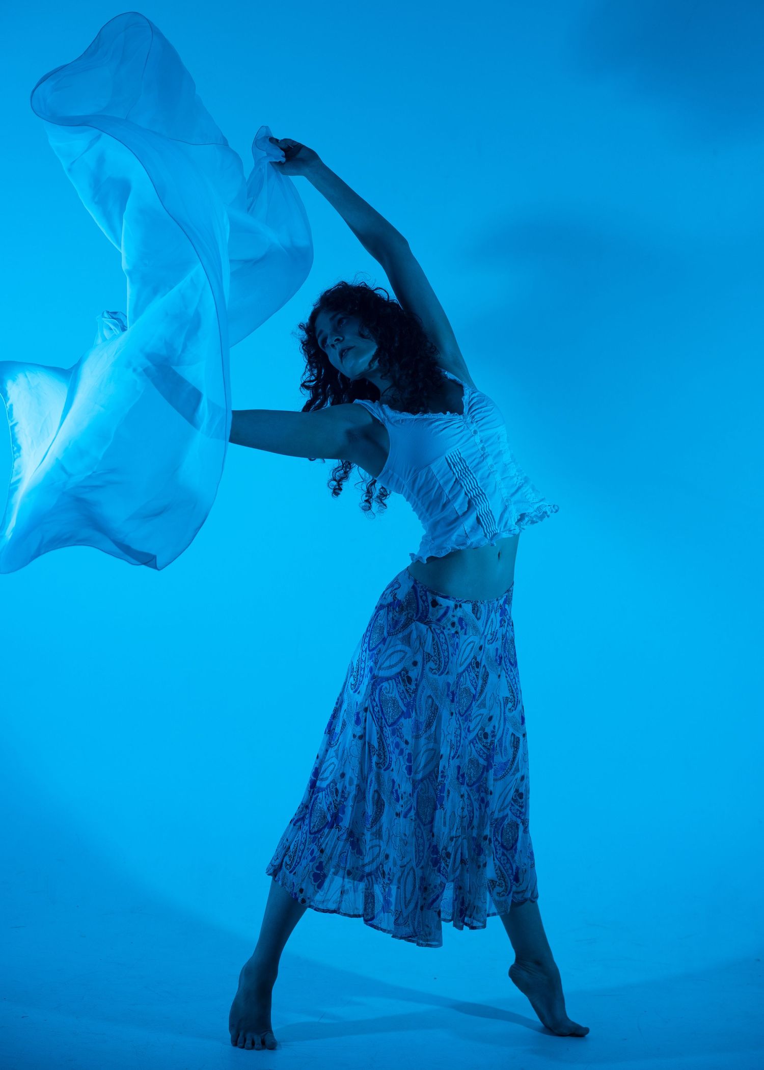 Woman dancing gracefully in a blue-lit studio, wearing a floral skirt and white top, waving a sheer fabric overhead.