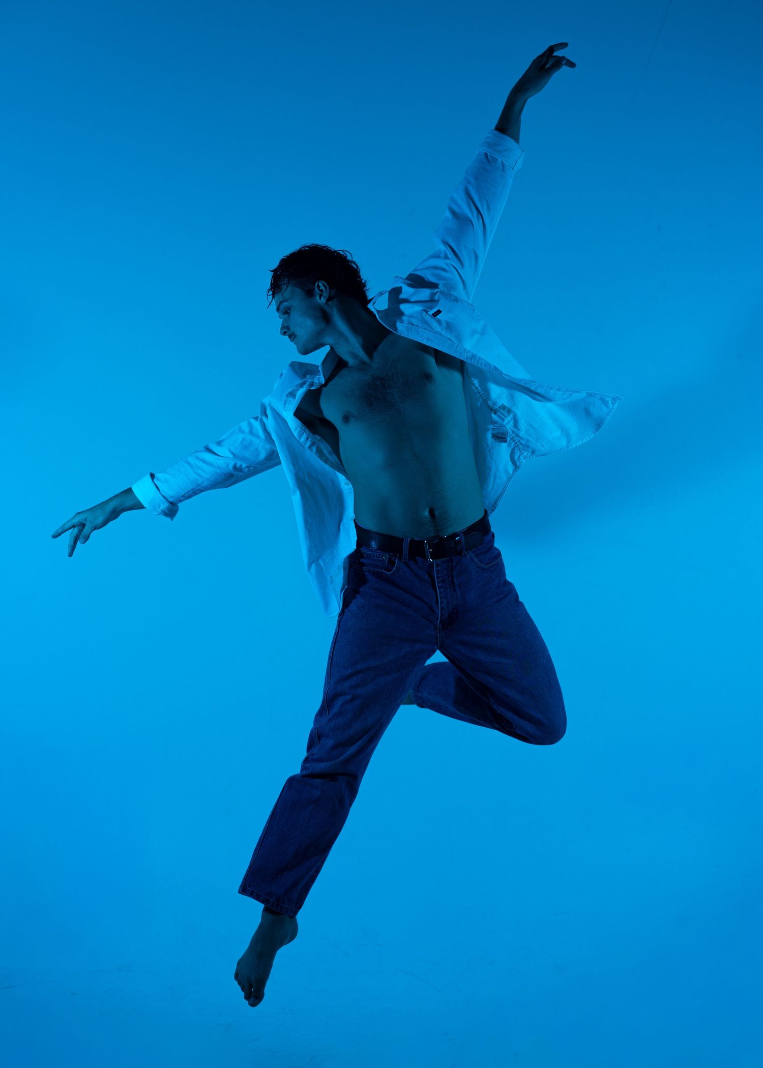 Male dancer leaps gracefully in an open white shirt and jeans against a vibrant blue background.