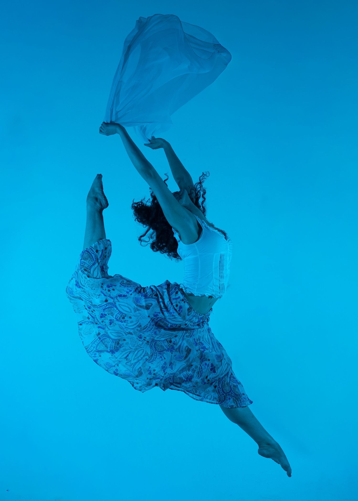 Dancer leaping gracefully against a blue background, holding flowing fabric in a dynamic aerial pose.