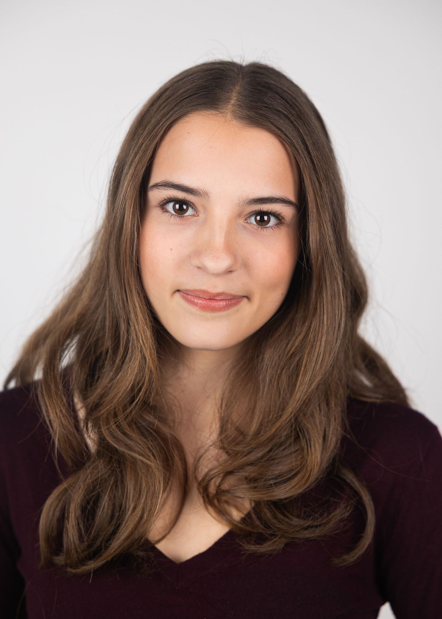 Young woman with long wavy brown hair wearing a dark maroon top against a white background.