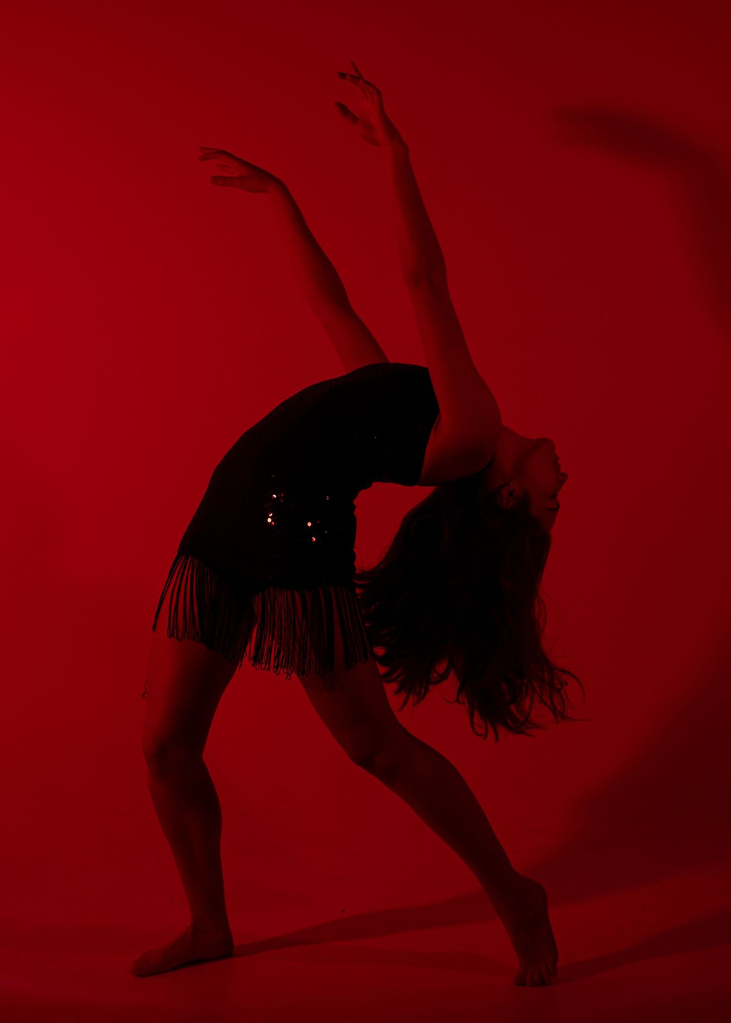 Dancer performs a dramatic backbend with arms extended in striking red-lit studio in black sequin outfit.