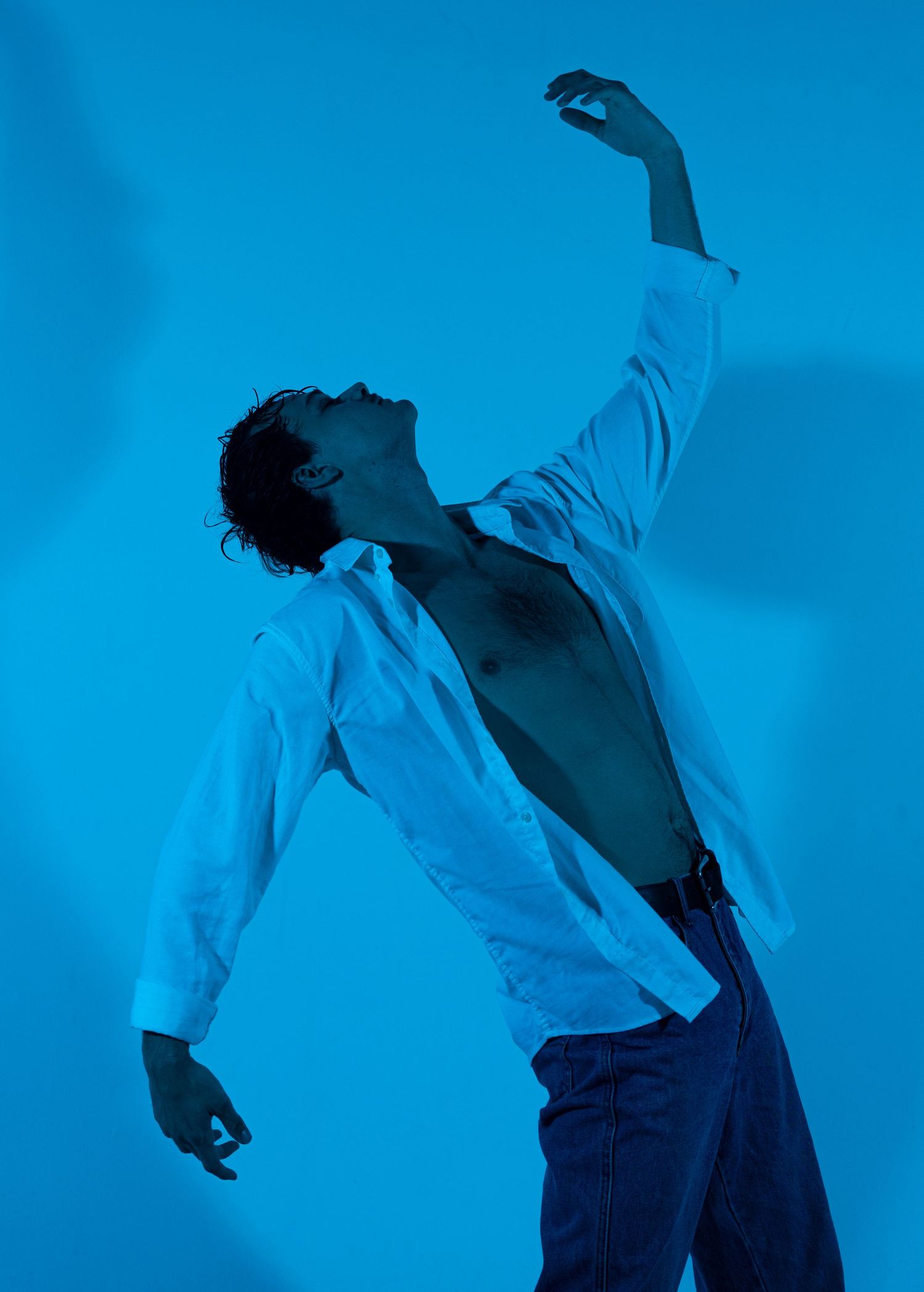 Man in open white shirt and jeans poses dramatically against a blue background with one arm raised.