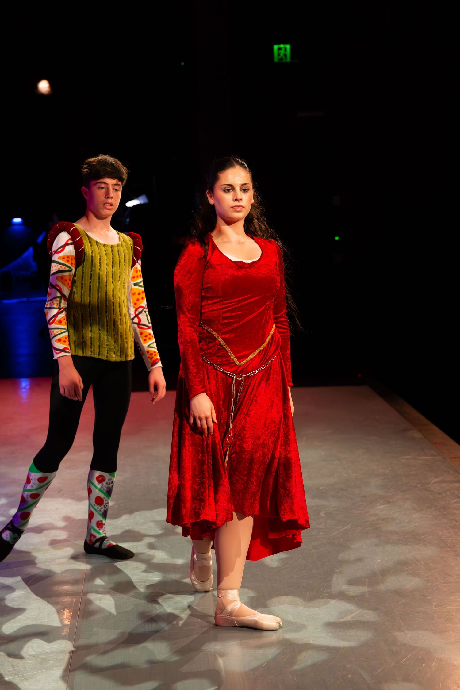 Two teen dancers in costume perform on stage, girl in red velvet dress and boy in colorful outfit.