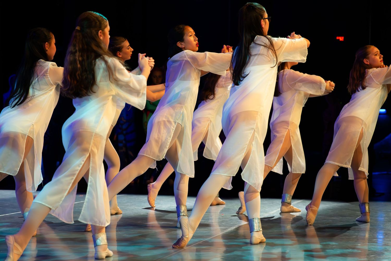 Group of young female dancers performing in white flowing costumes on a stage under colorful lights.