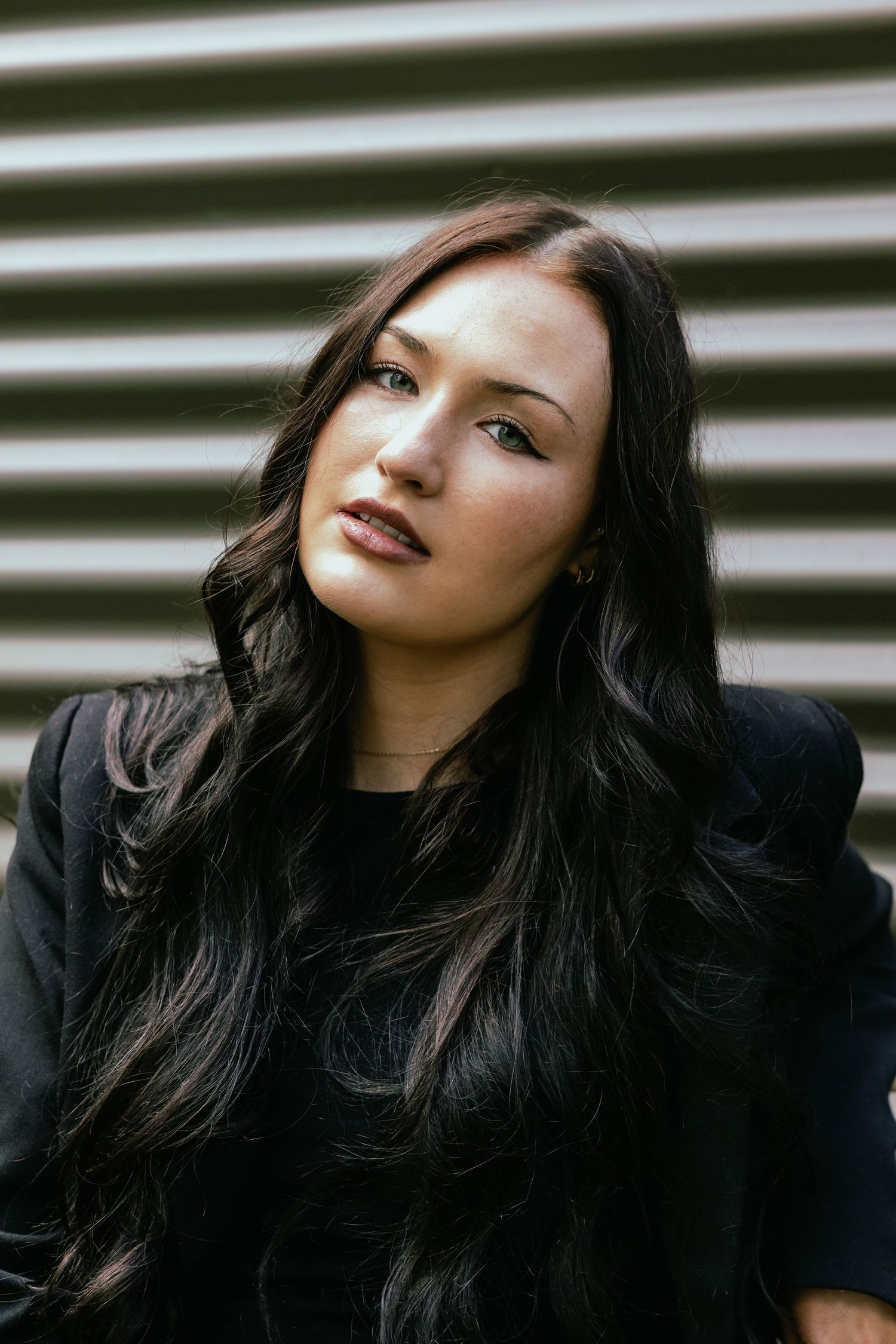 Young woman with long dark hair wearing black outfit posing against striped metal wall.