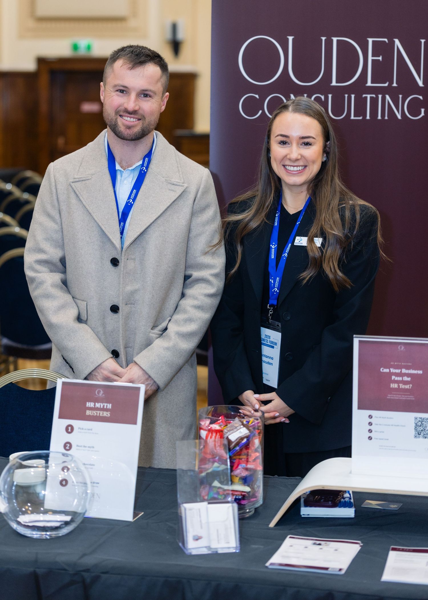 Two professionals stand at anOuden Consulting booth at a networking event, smiling beside a display table.