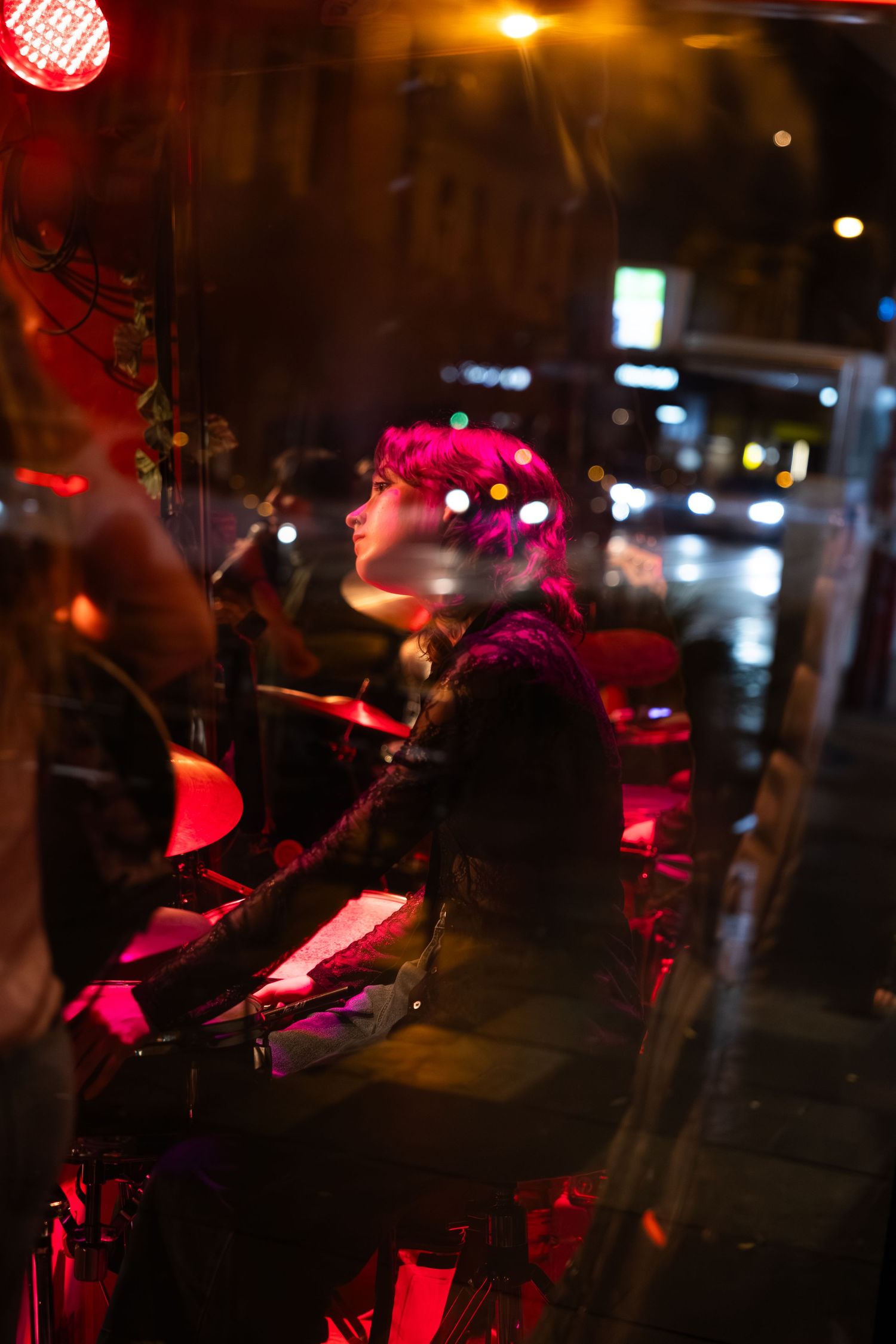 A musician performs behind a drum set in a dark venue illuminated by pink and red stage lights.
