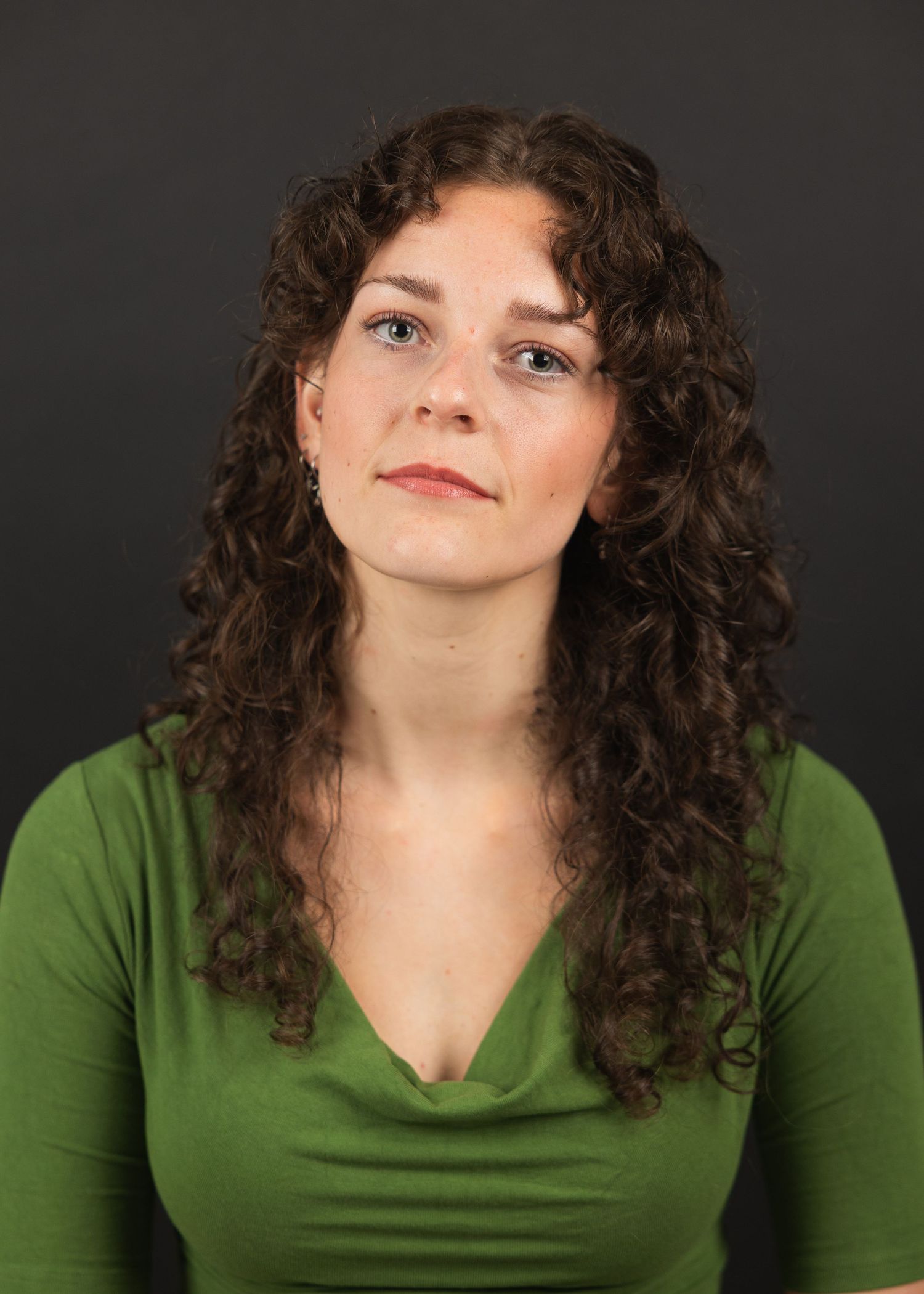 Woman with curly brown hair wearing a green v-neck top, posing against a dark gray background.