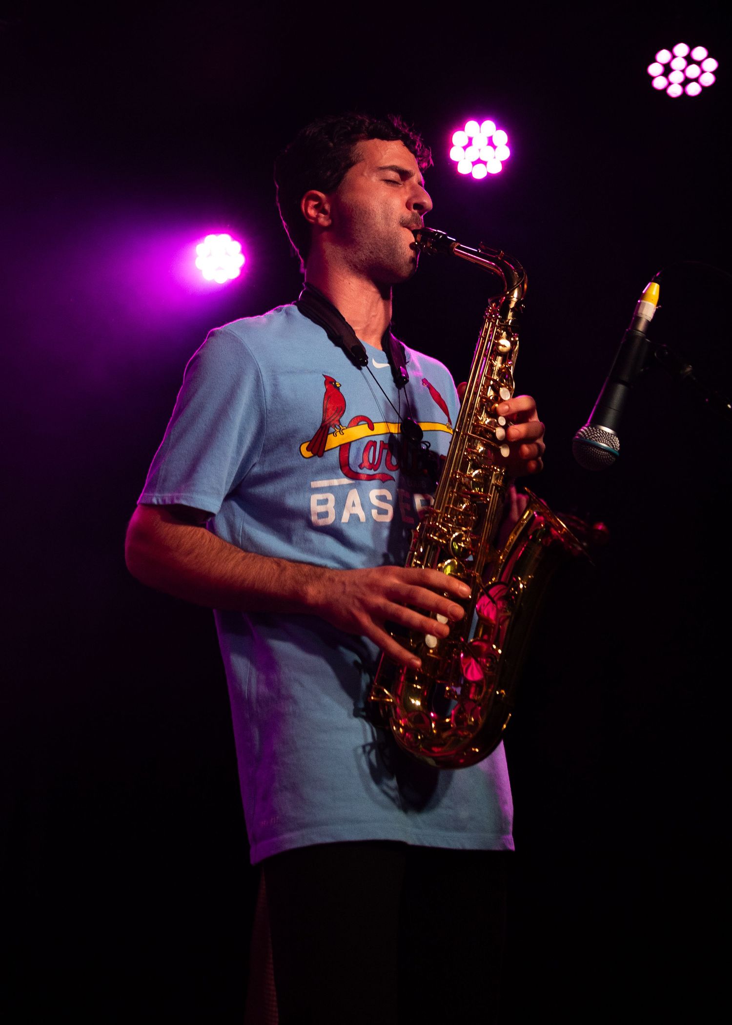 Young man playing alto saxophone on stage, wearing a St. Louis Cardinals baseball jersey under pink stage lights.