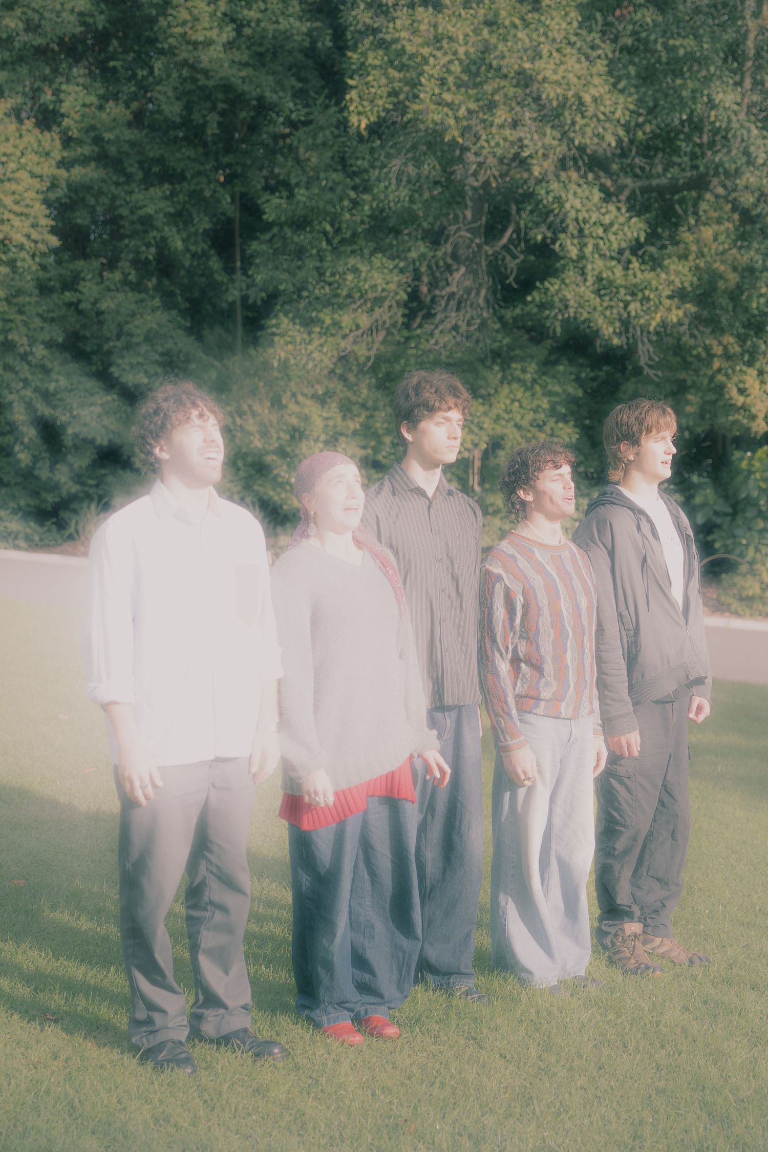Five young people standing in a row on grass with green trees behind them.