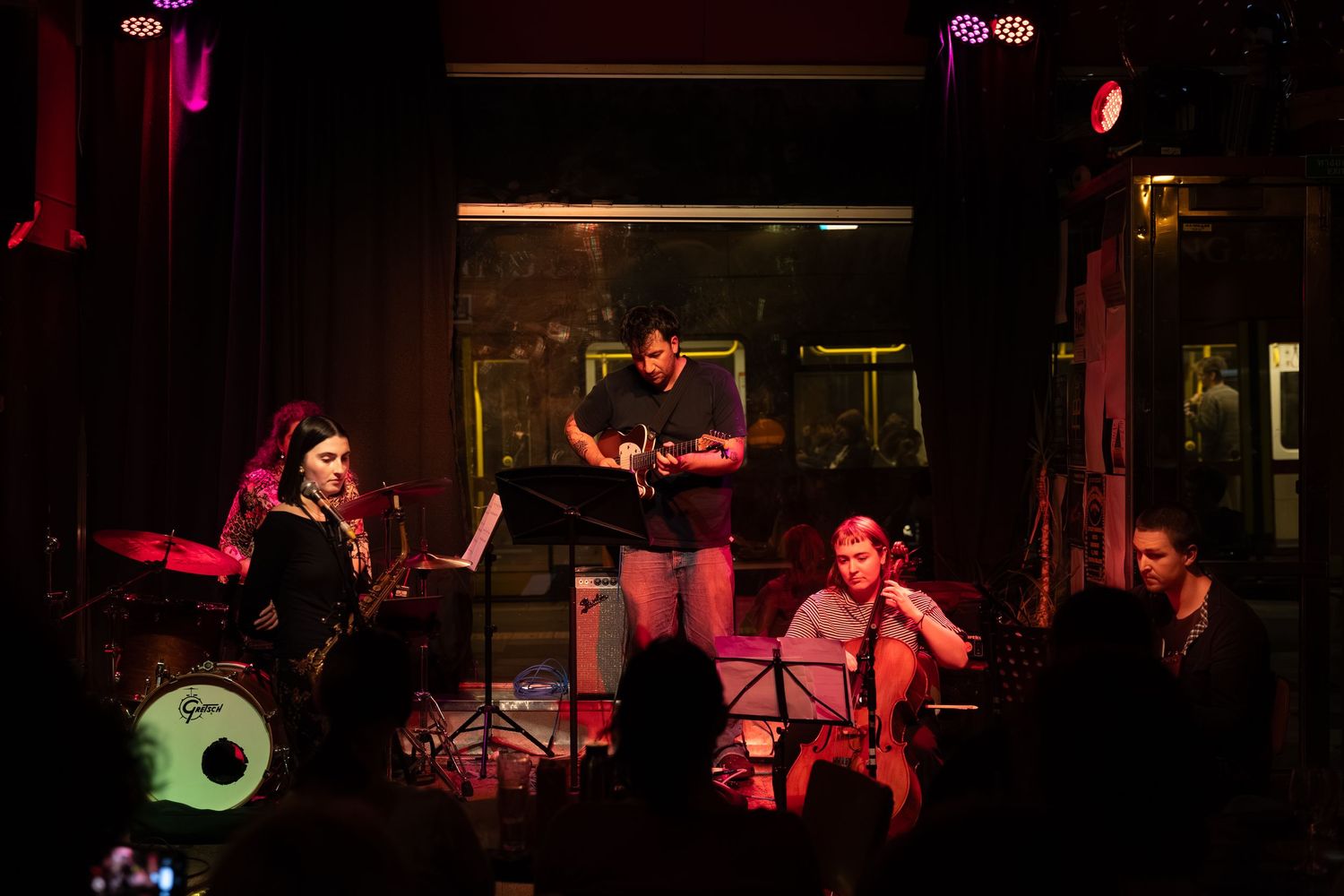 Musicians perform on stage with instruments under dramatic red and green lighting at a live music venue.