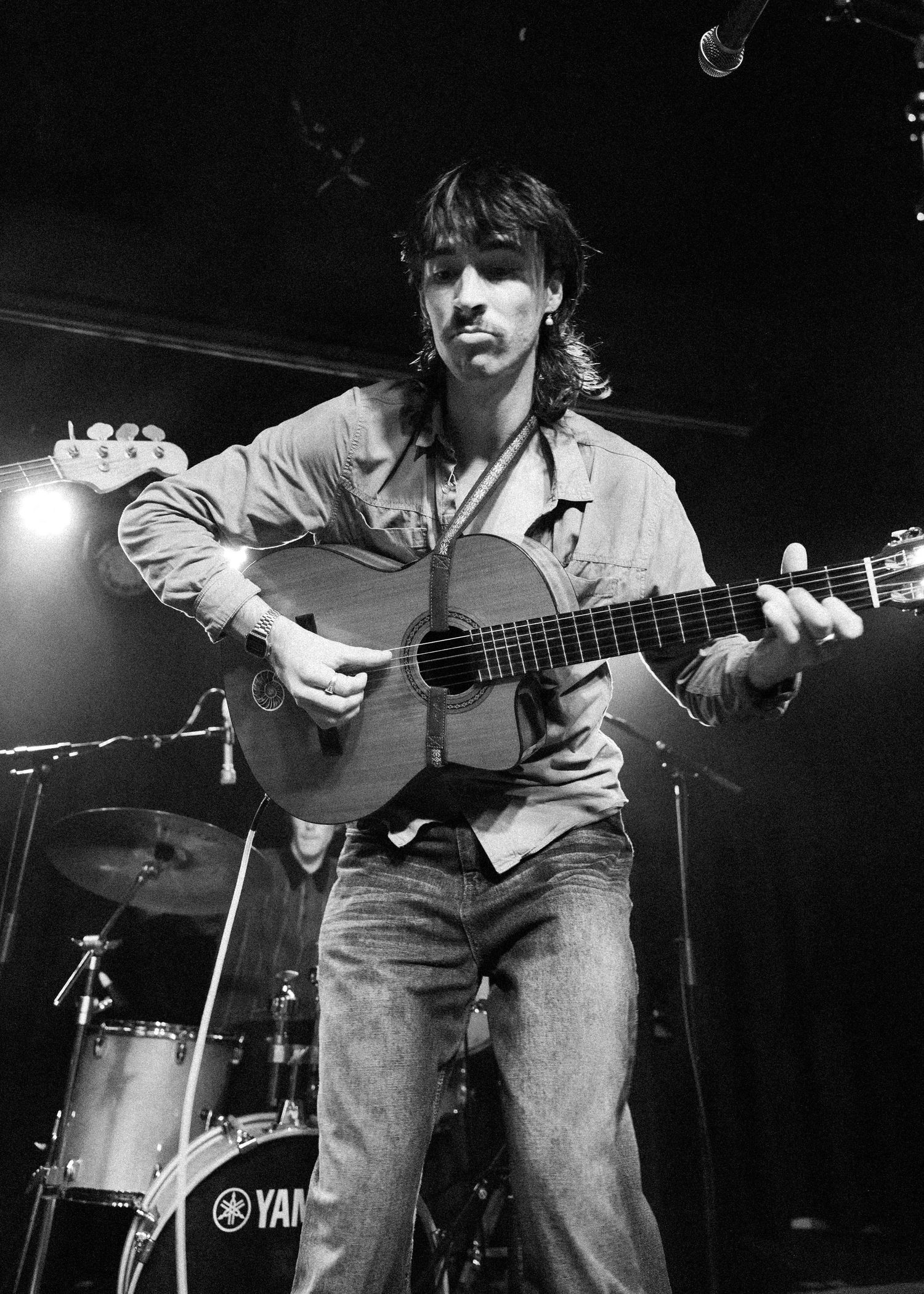 Black and white photo of a long-haired male guitarist playing acoustic guitar on stage with a Yamaha drum kit behind him.