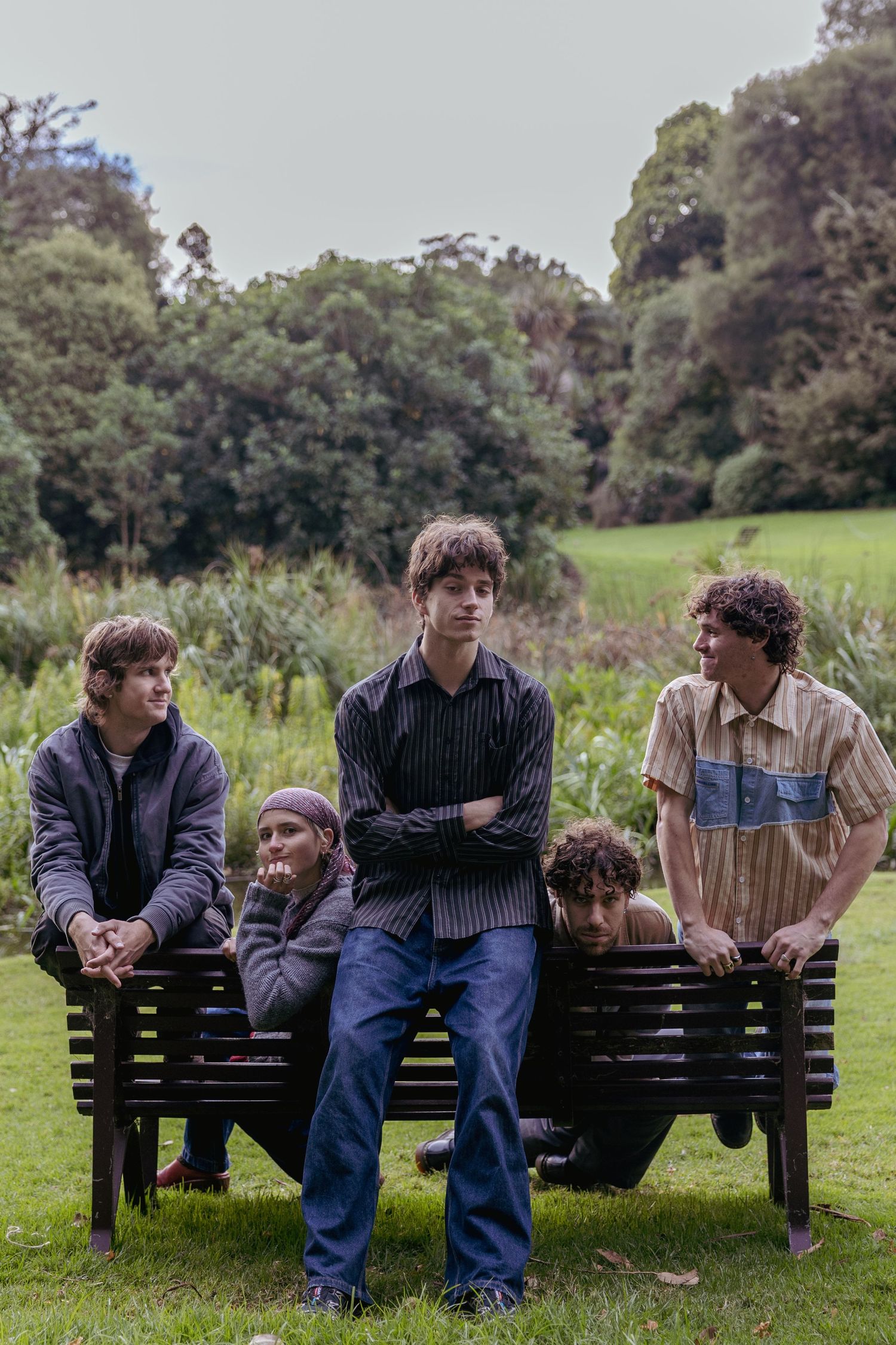 Five casually dressed young people pose on and around a wooden park bench on green grass.