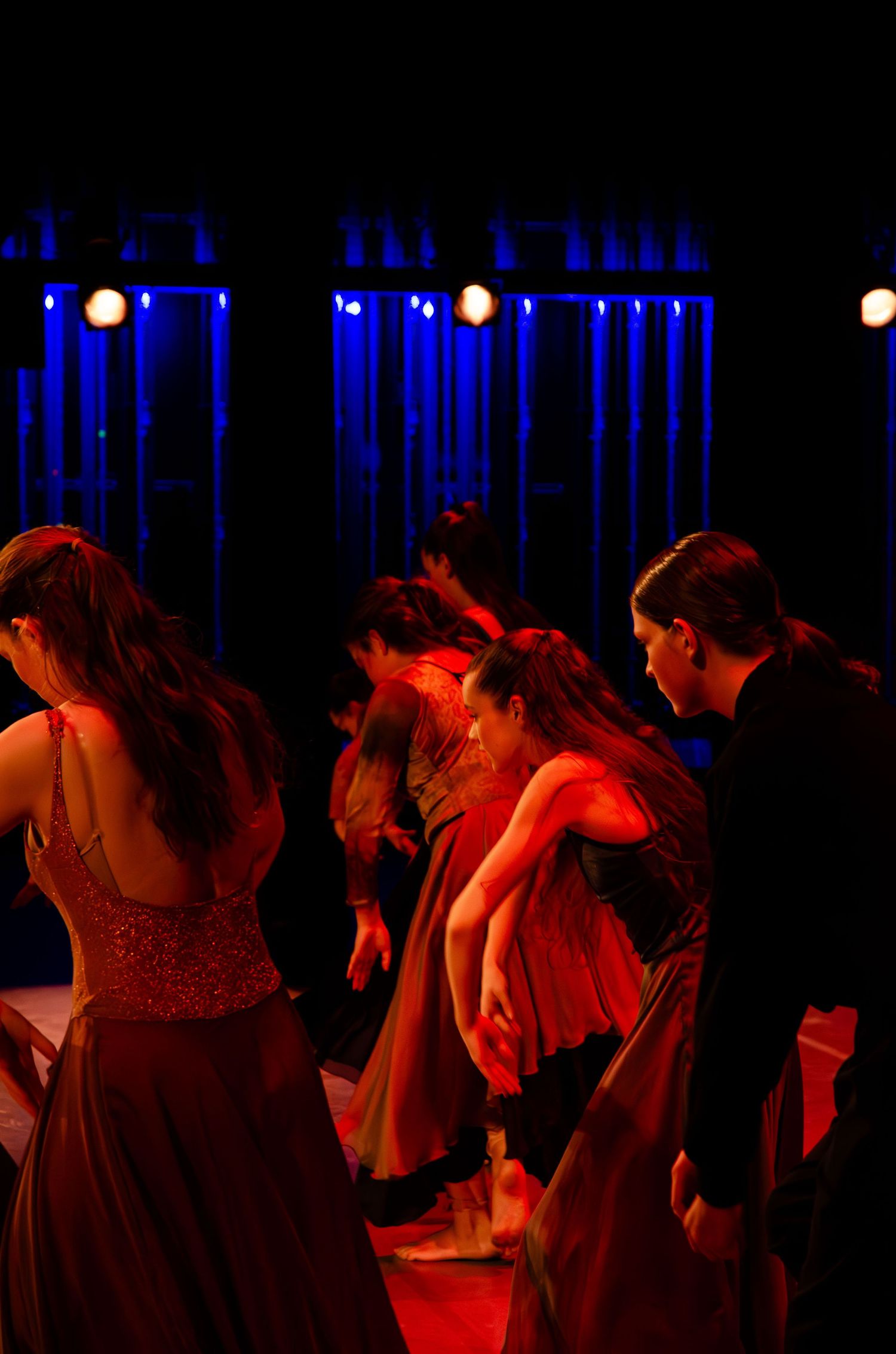 Dancers in red gowns perform on a dramatic stage lit with blue and amber lights during an evening performance.