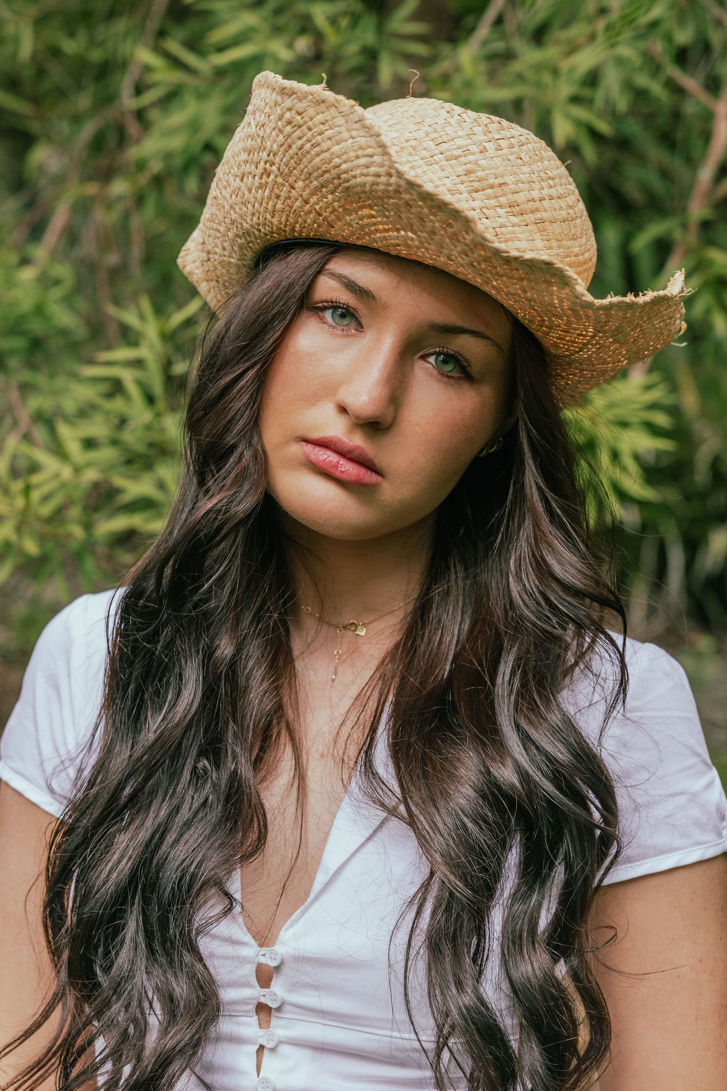 Young woman with long dark hair wearing a straw cowboy hat and white top, posing outdoors near green foliage.