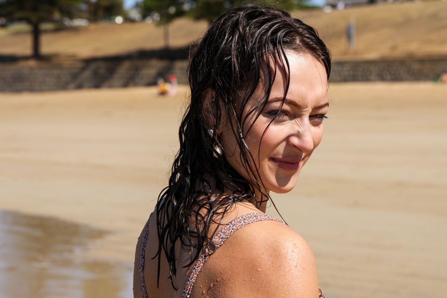 Smiling woman with wet dark hair at a sunny beach, wearing a swimsuit, looking over her shoulder.