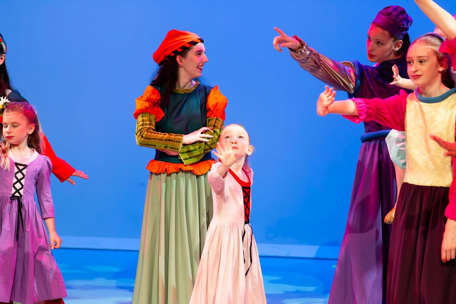 Children perform in a colorful stage production, wearing medieval costumes on a blue backdrop.