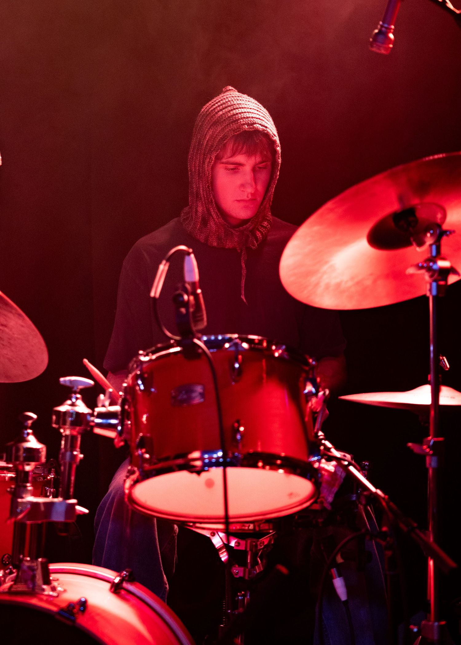 Young drummer in a hooded sweater plays a red drum kit on stage under dramatic red lighting.