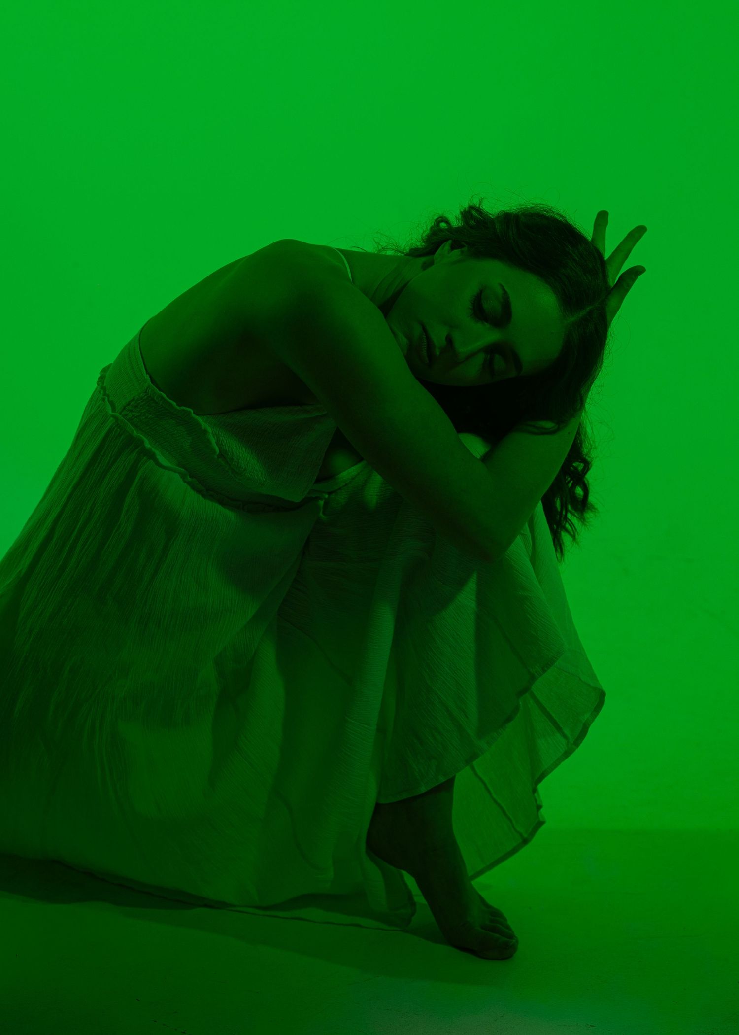 A dancer in a white dress crouches gracefully under dramatic green studio lighting.