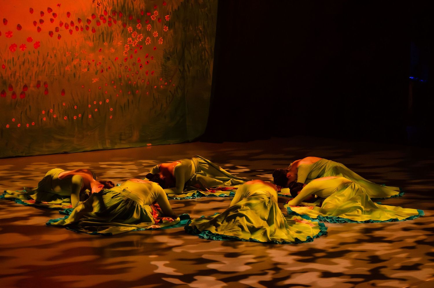 A series of dancers in colorful costumes perform a contemporary dance piece on a dark stage with dramatic lighting.