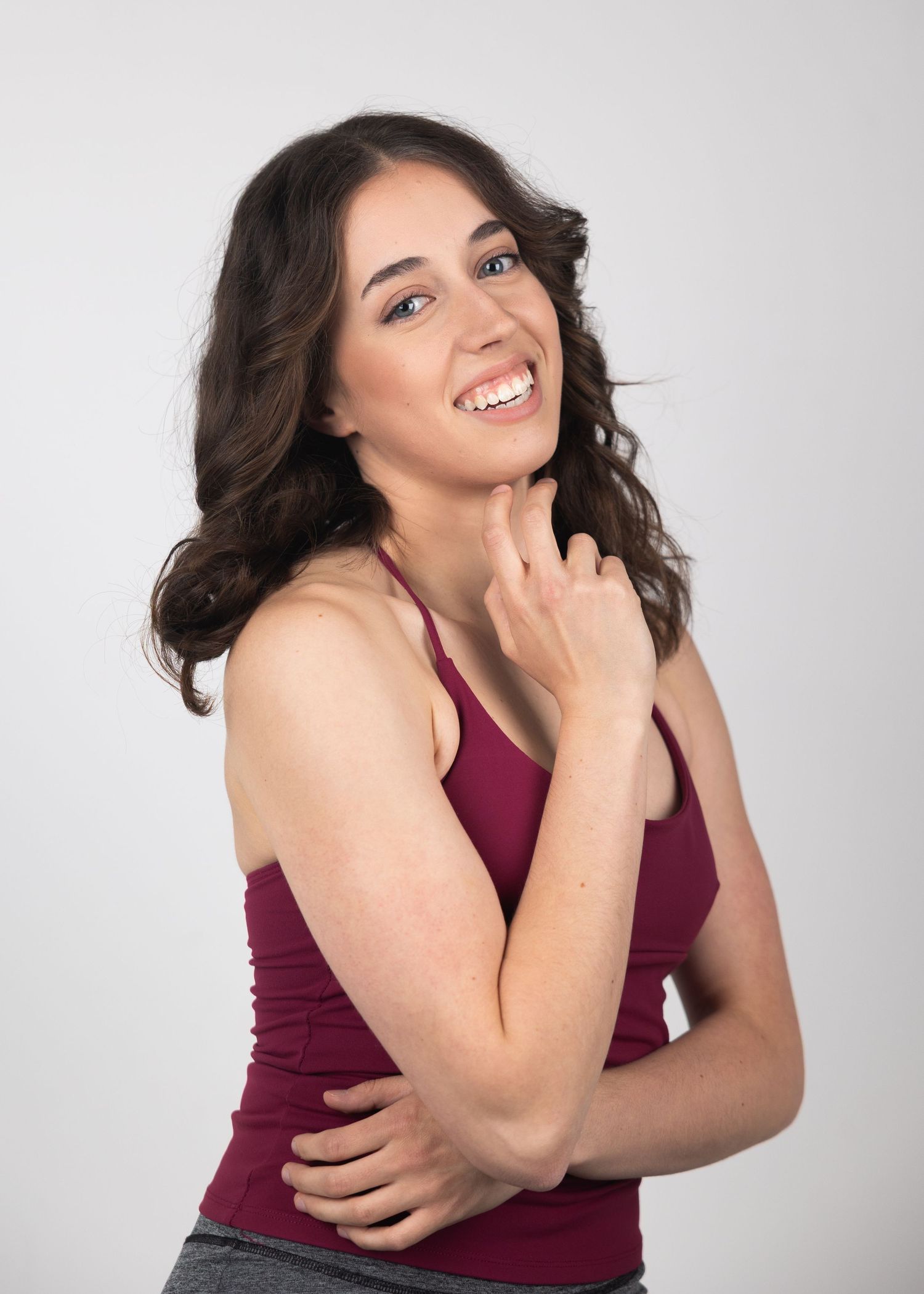 Smiling young woman with dark wavy hair wearing a burgundy tank top posing confidently against a white background.