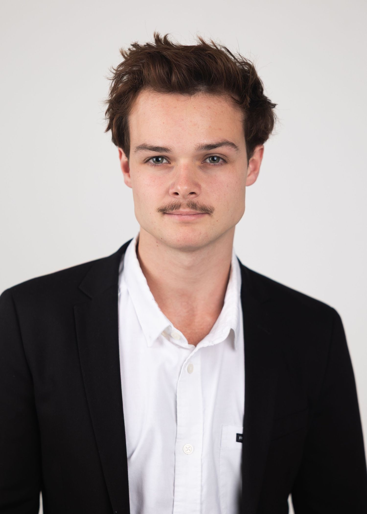 Young man with mustache wearing black blazer and white shirt poses for professional headshot photo.